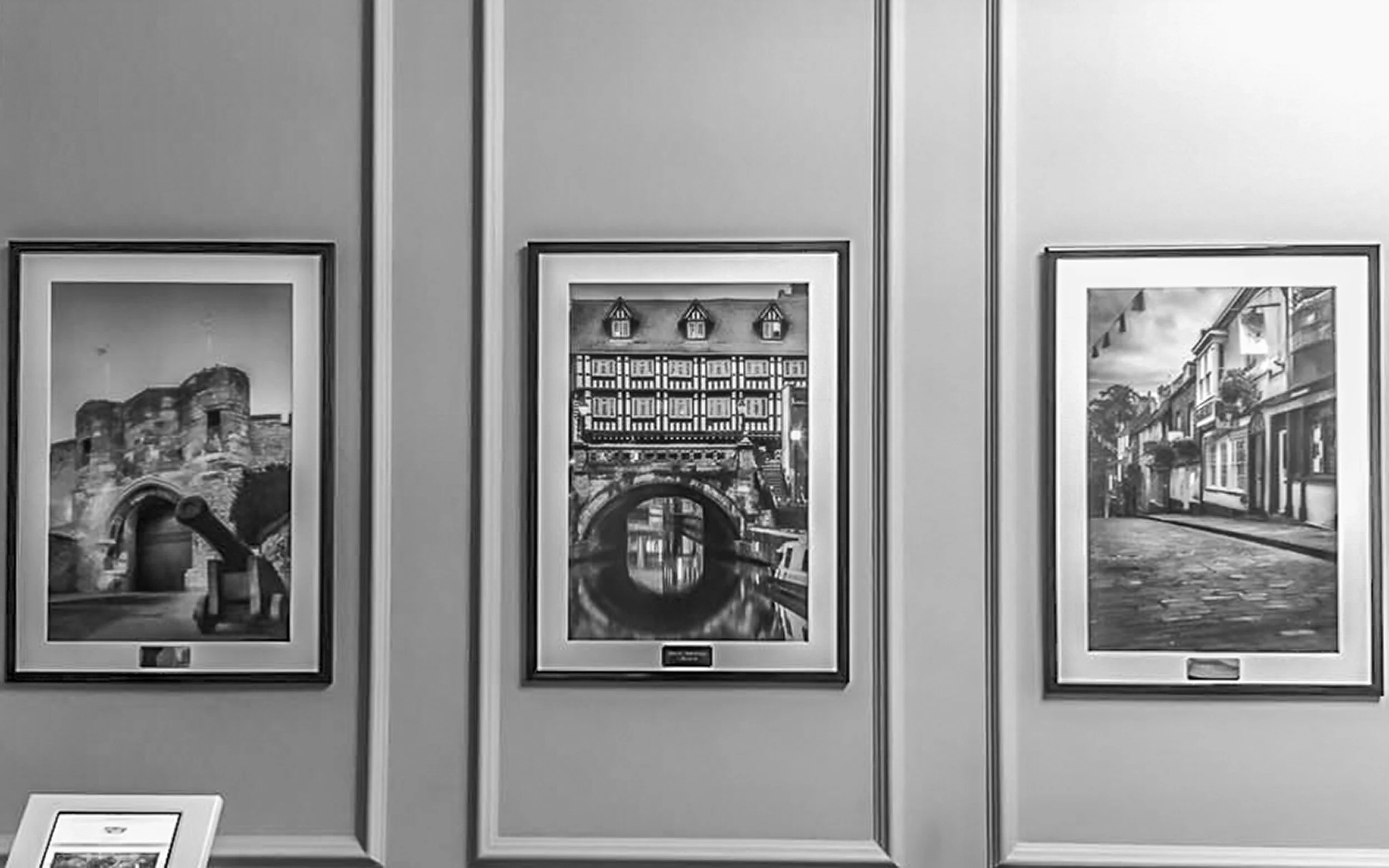 Black and white photograph of an art gallery wall displaying three framed pictures of European city scenes.