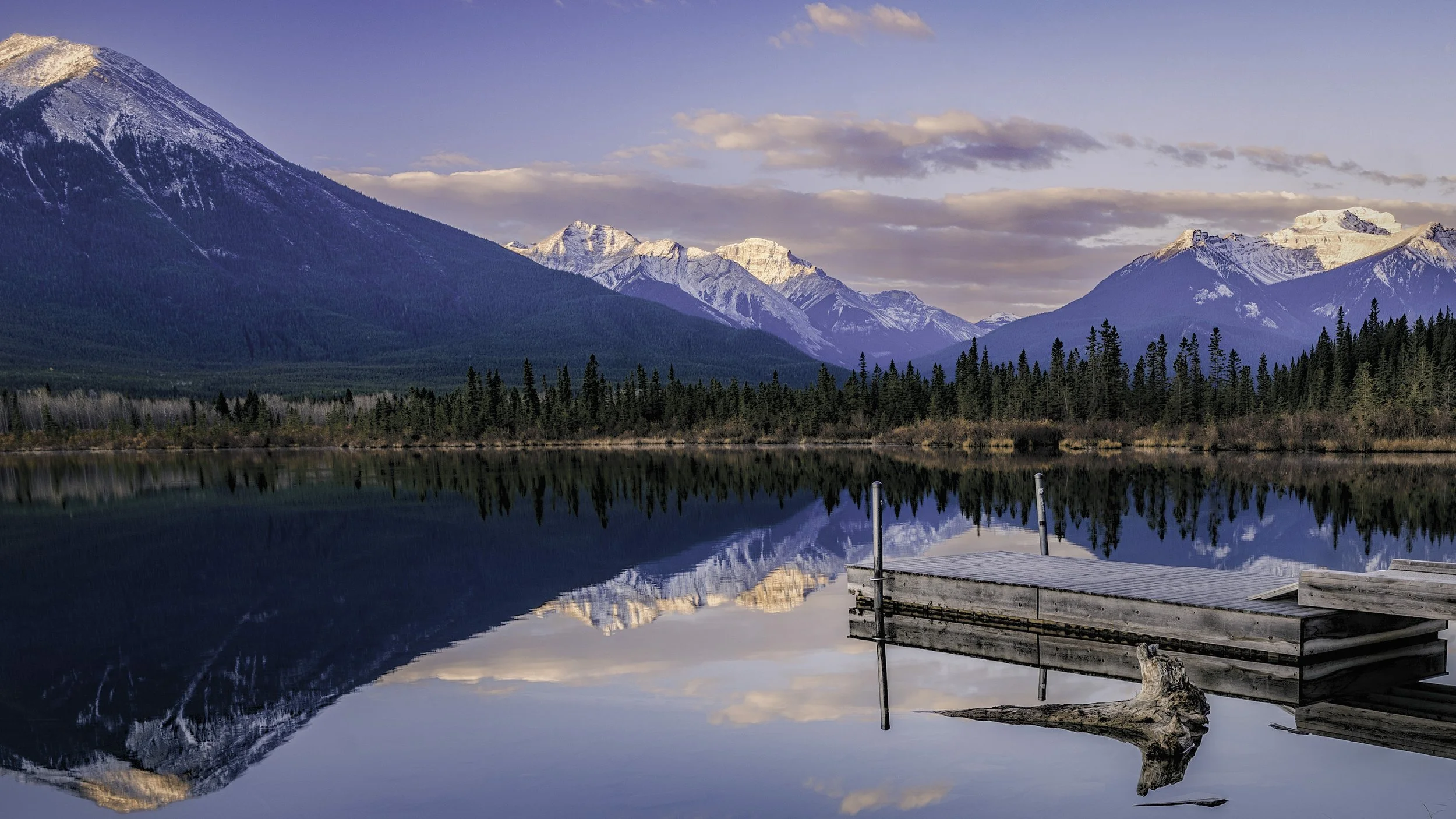 A serene mountain lake reflecting snow-capped peaks, dense pine trees, and a partly cloudy sky, with a wooden dock and a piece of driftwood in the water.