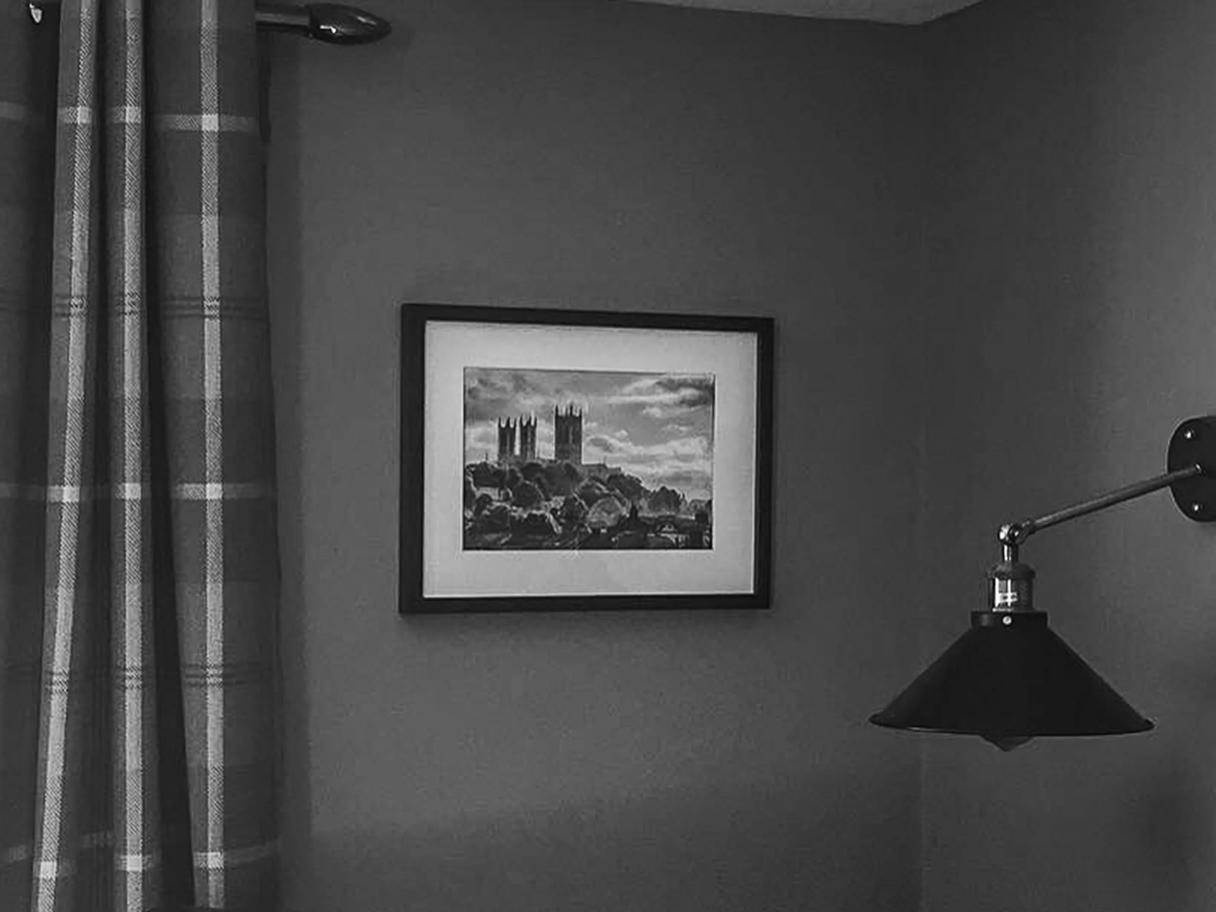 A framed painting of a castle on a hilltop hangs on a plain wall, with a curtain to the left and a modern lamp with a black shade to the right.