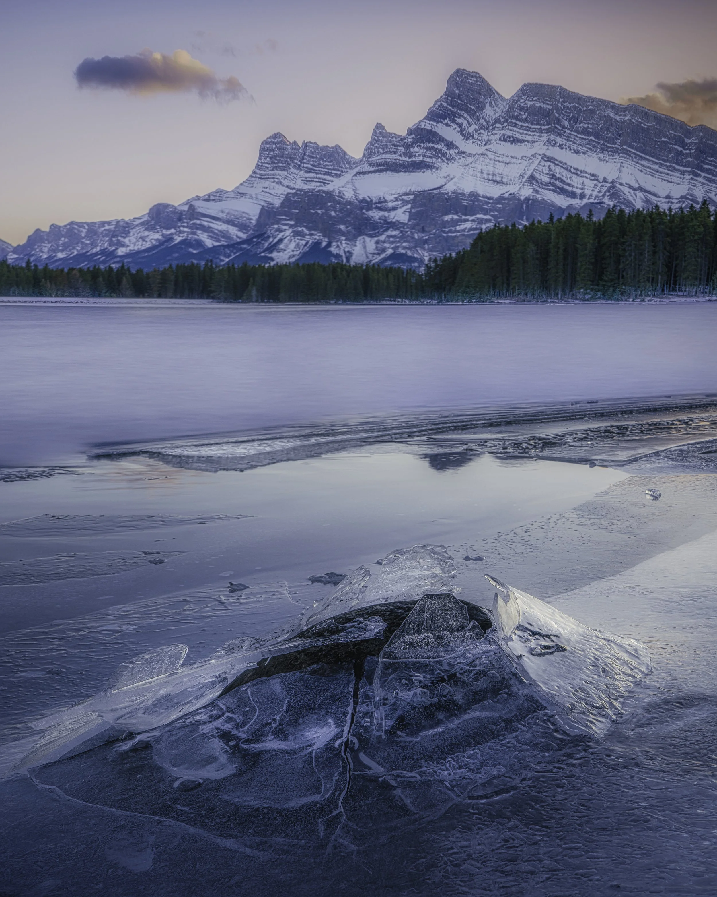 Snow-covered mountains in the background, a forest along the shoreline, and a frozen lake with ice formations in the foreground.