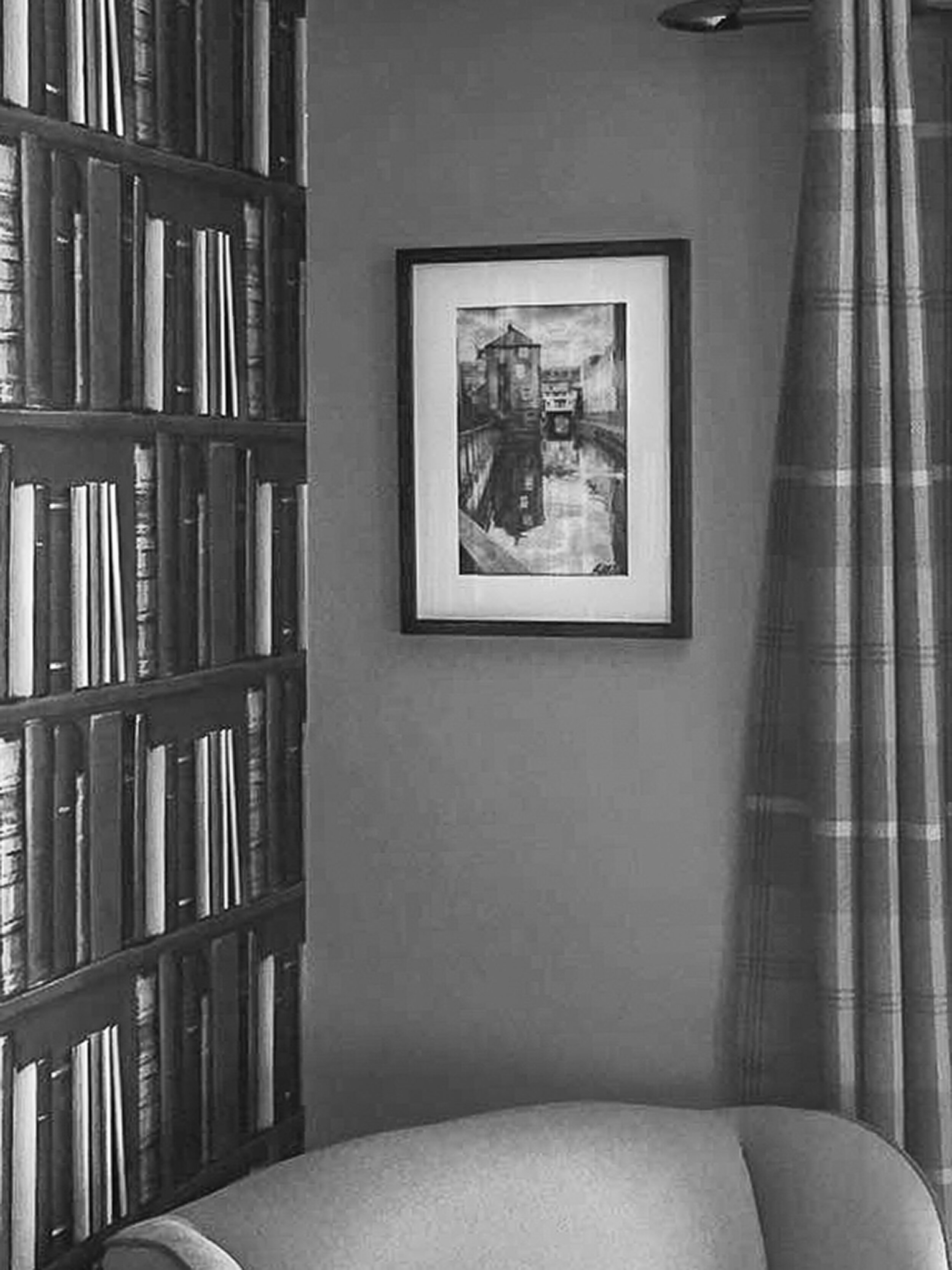 A black and white photo of a corner of a room with a wooden bookshelf filled with books on the left, a framed watercolor painting of a building reflected in a canal on the wall, and a curtain with plaid pattern on the right, next to a piece of furnit