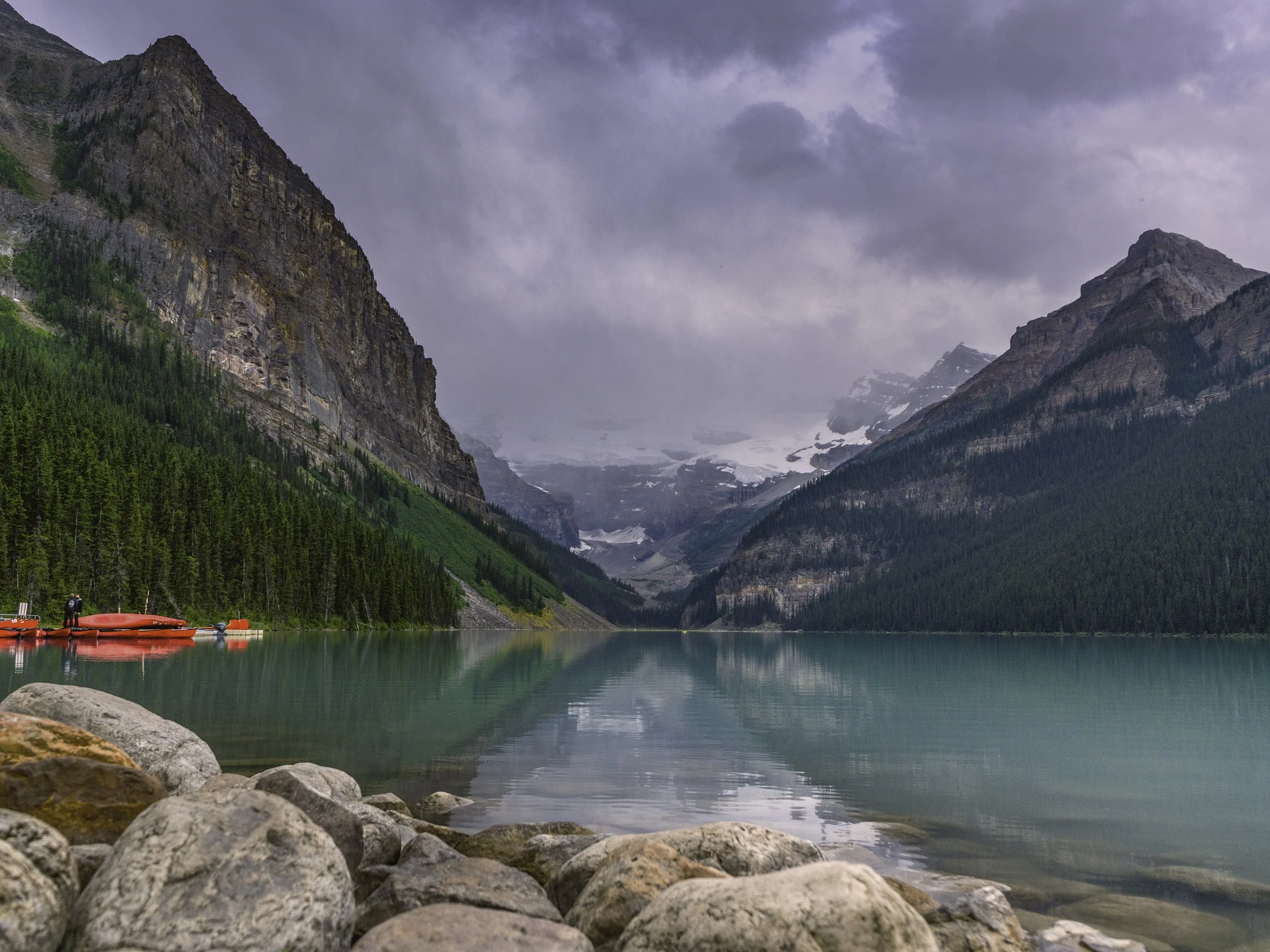 A scenic view of a mountain lake with green pine trees on the slopes, snow-capped mountains in the background, and a cloudy sky overhead. Red canoes are docked along the rocky shore.