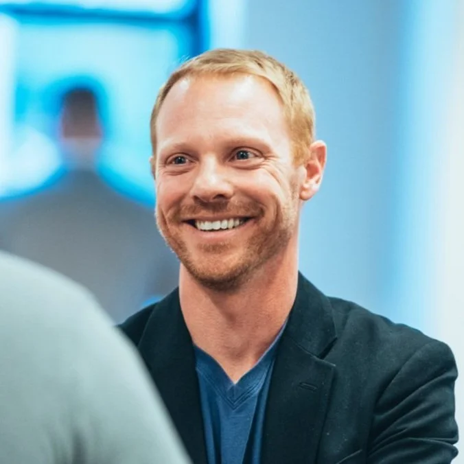 Headshot of Billy Craig. White man with reddish blonde hair standing and smiling talking to a crowd.