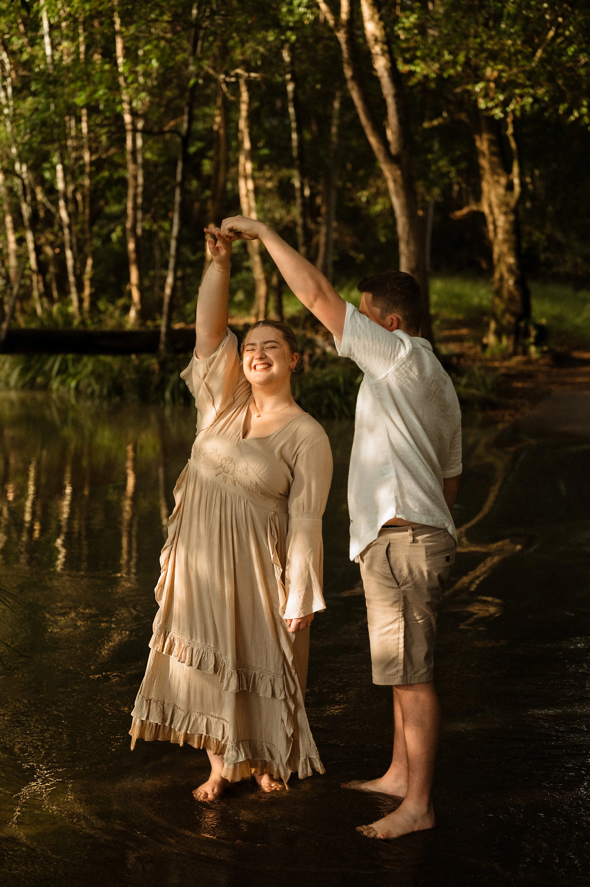 A young woman in a beige dress and a young man in a white shirt and shorts dancing barefoot in a forested area near a creek, with sunlight filtering through trees.