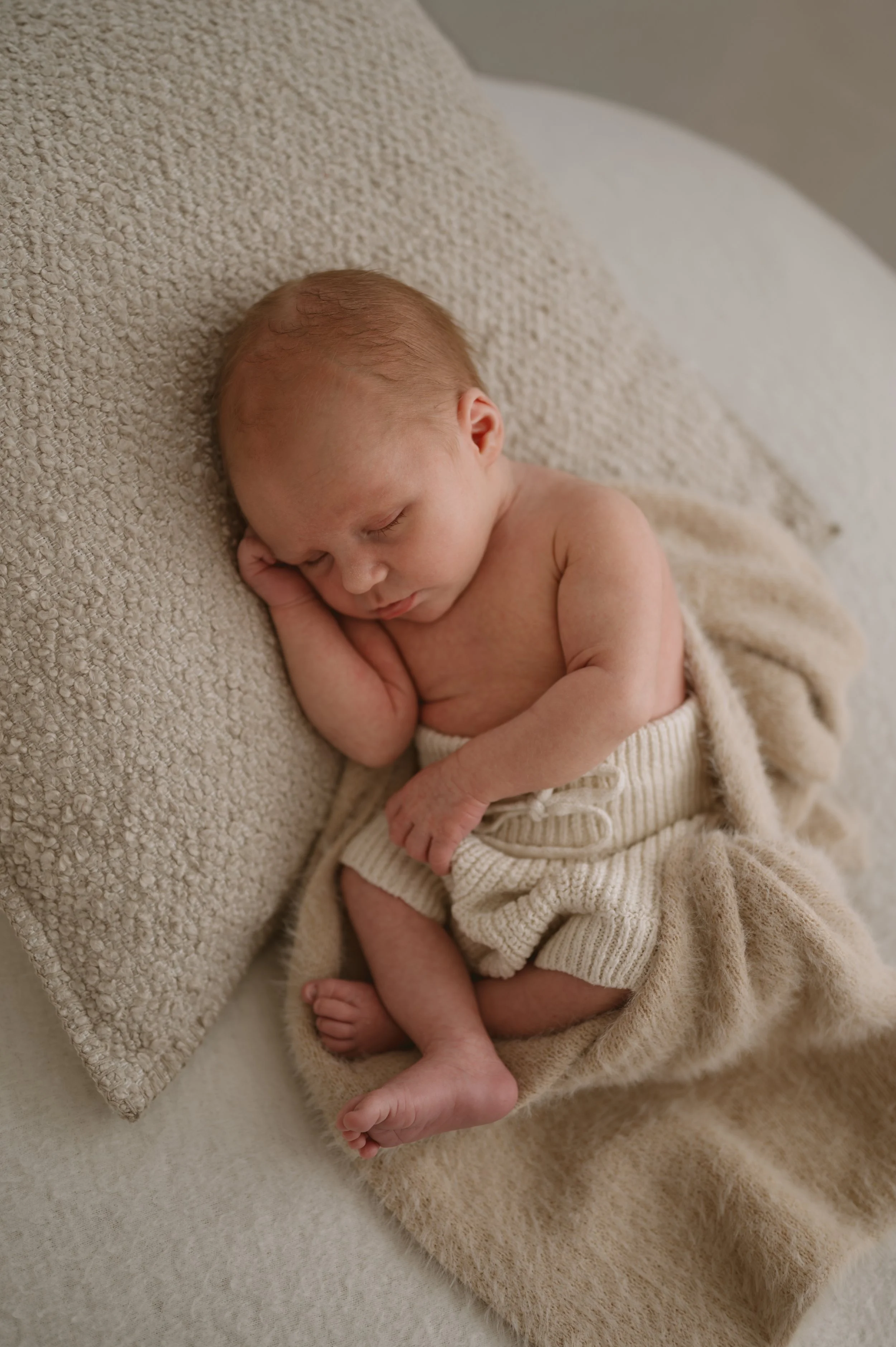 A sleeping baby lying on a cream-colored textured blanket with a matching pillow, dressed in cream knit shorts, peacefully resting with one hand under the cheek.