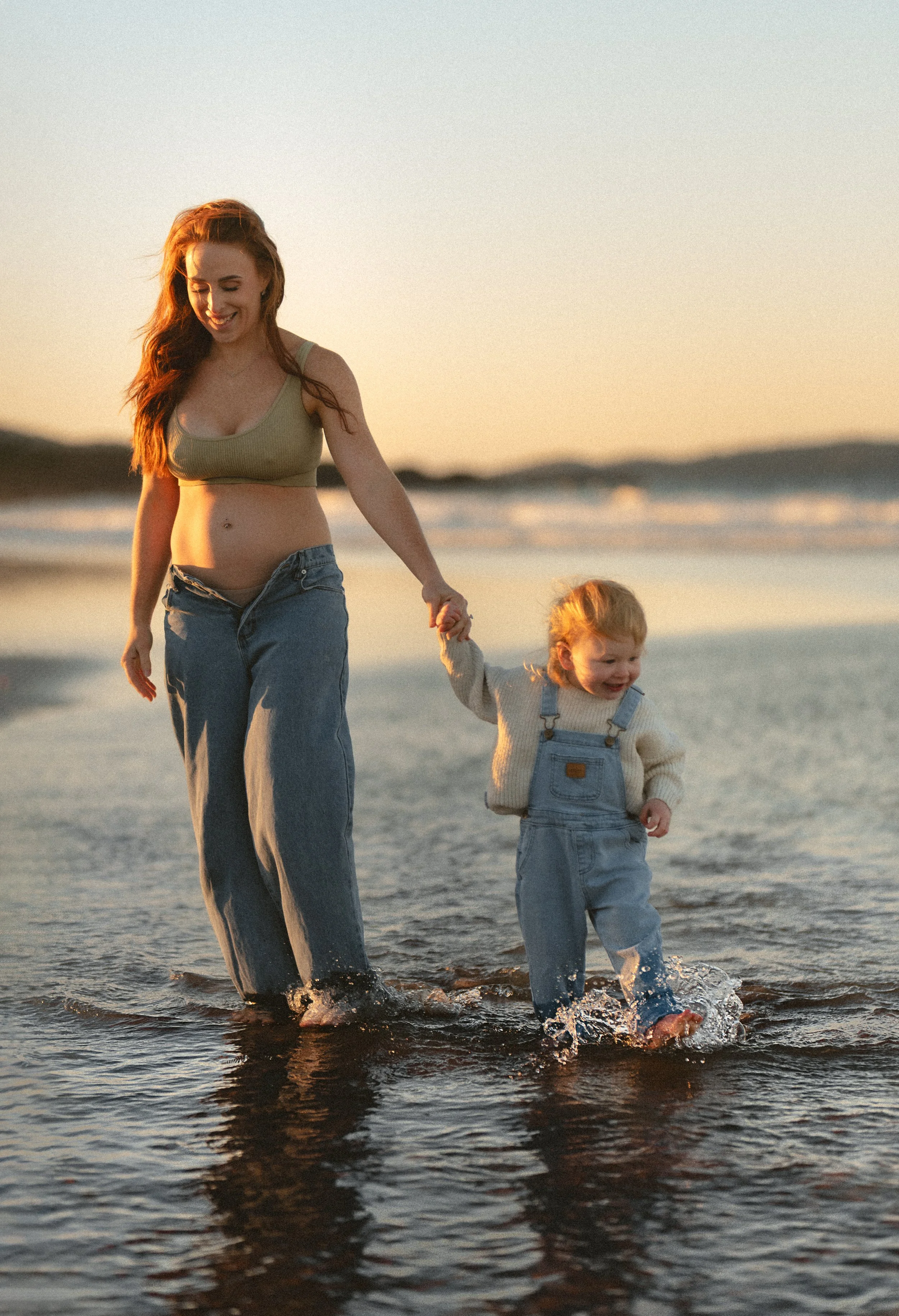 A woman and a young girl walking hand-in-hand through the shallow water at the beach during sunset.