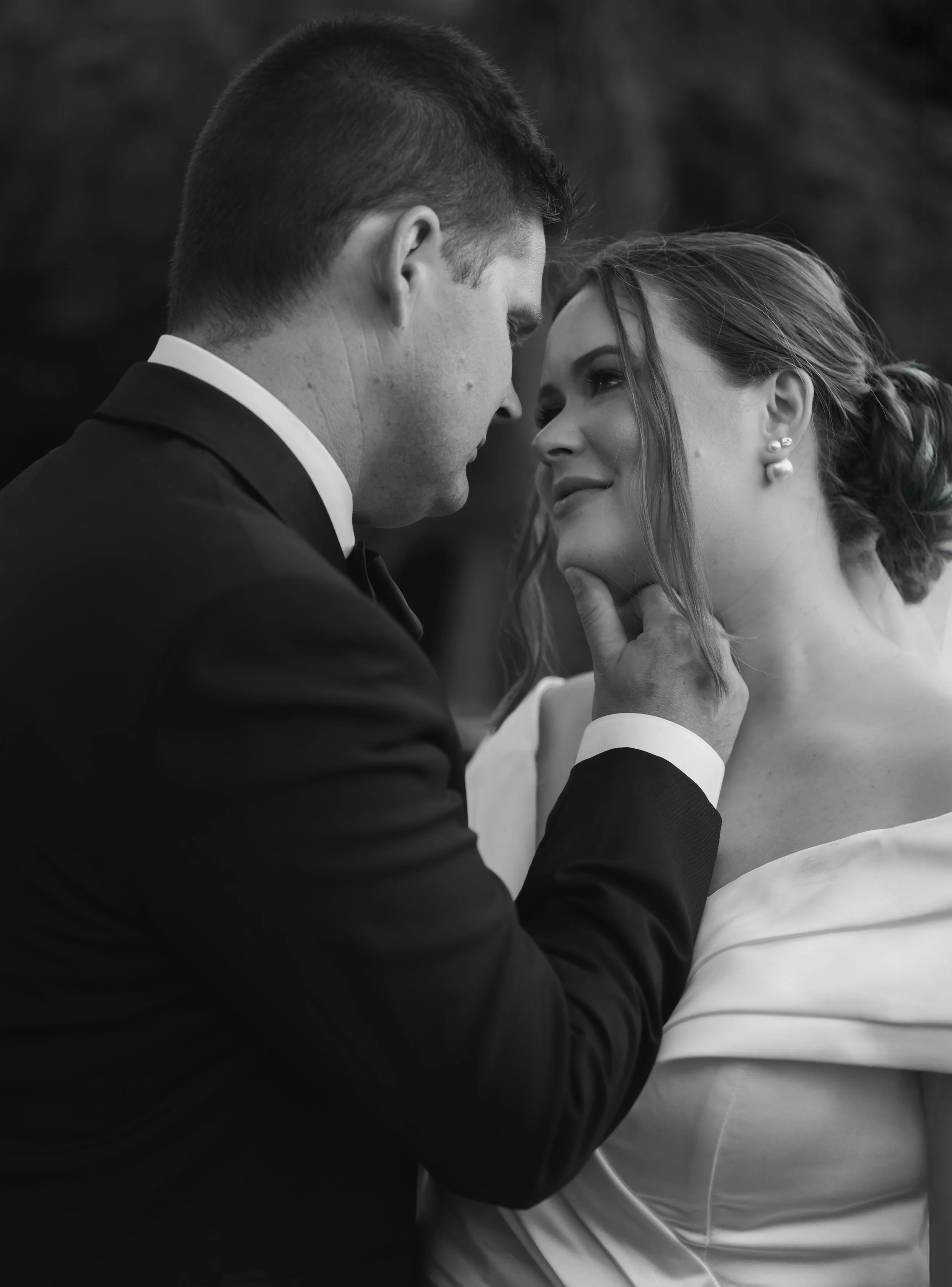 A black and white photo of a man and woman in an intimate moment. The man is gently holding the woman's face, and they are looking into each other's eyes. The woman is smiling softly, with her hair styled in an updo and wearing earrings.