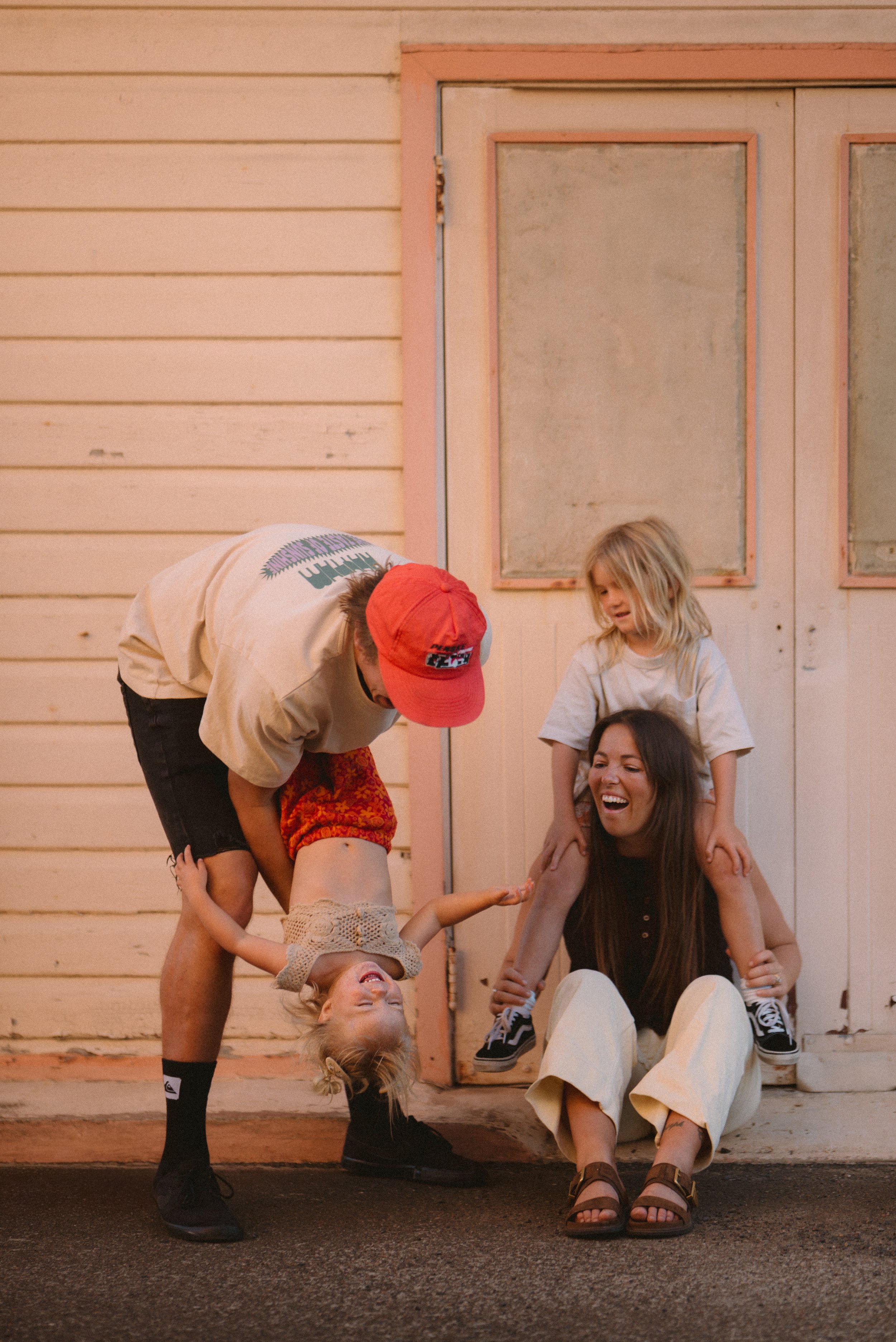 A family of four playing and laughing outside a house with light wood siding and a pink door. The mother is sitting on the ground with a girl on her shoulders, while the father is holding the girl upside down.