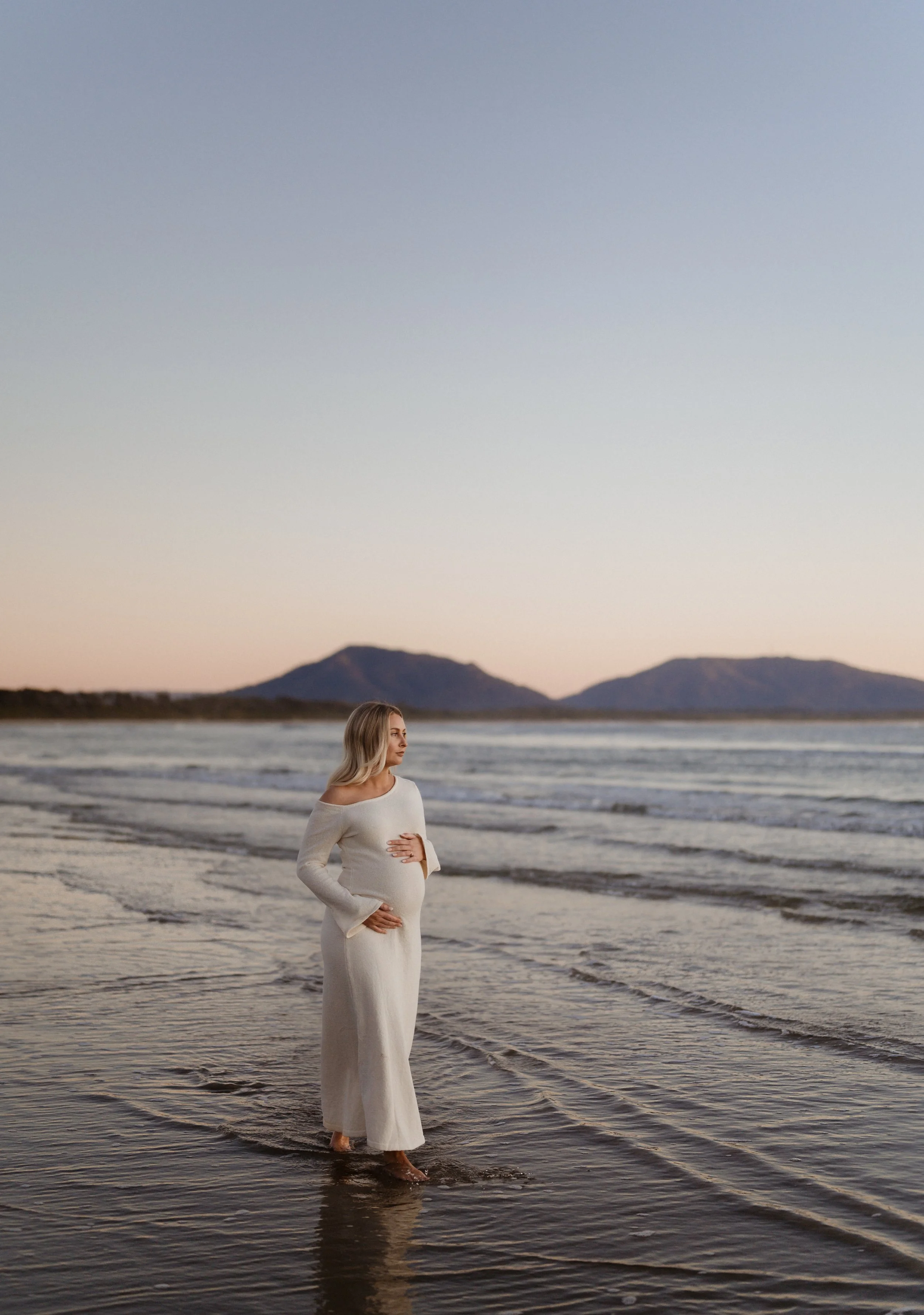 A pregnant woman in a white dress standing ankle-deep in the water at the beach during sunrise, with mountains in the background.