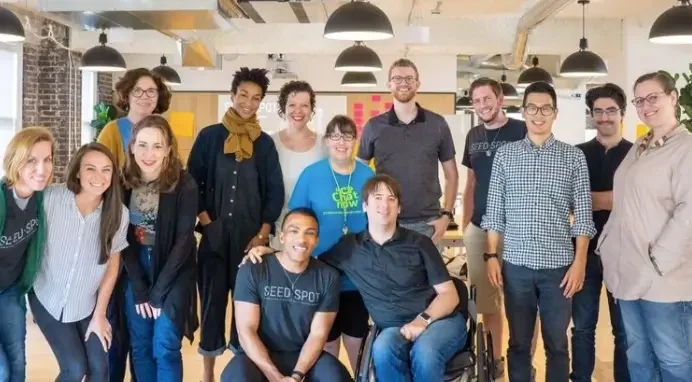 Group of twelve diverse people smiling in an open office space, some standing, some sitting, with hanging lights and casual decor.