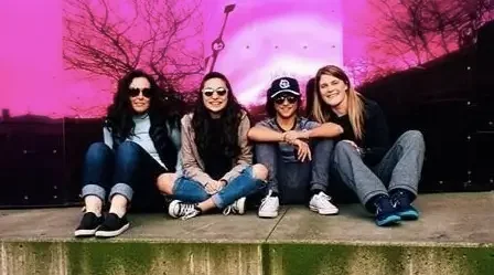 Four women sitting on the ground in front of a reflective pink wall, smiling at the camera, with leafless trees reflected in the wall.