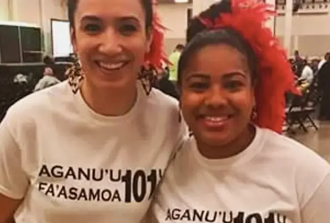Two women smiling and wearing matching white t-shirts with Hawaiian text, one with red feathered hair and the other with leopard print earrings, in a crowded indoor setting.