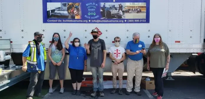 Group of seven people standing in front of a large white trailer with a banner, some wearing masks, the person second from the right is looking at their phone, and the person on the far left is wearing a reflective safety vest.