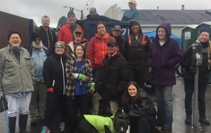Group of people standing outdoors in front of a large truck, dressed in casual and outdoor clothing, with some wearing gloves, hats, and jackets, posing for a photo with a black dog wearing a green vest.