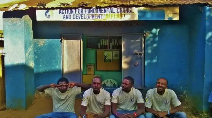 Four young men sitting outside a blue building with open doors, wearing matching white T-shirts, with a banner above them that reads 'Action for Fundamental Change and Development.'