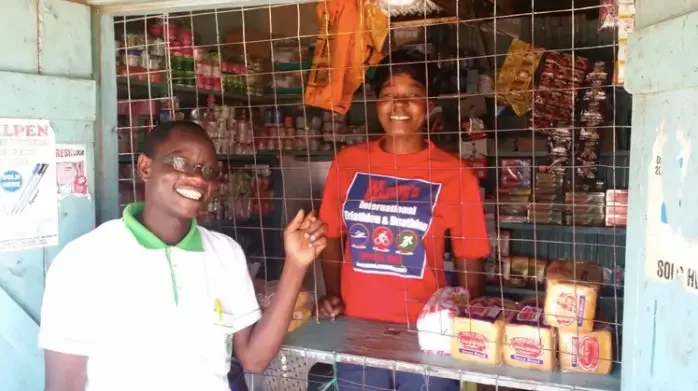 Two women smiling inside a small shop with products behind wire mesh, one in a white shirt and glasses, the other in a red shirt.