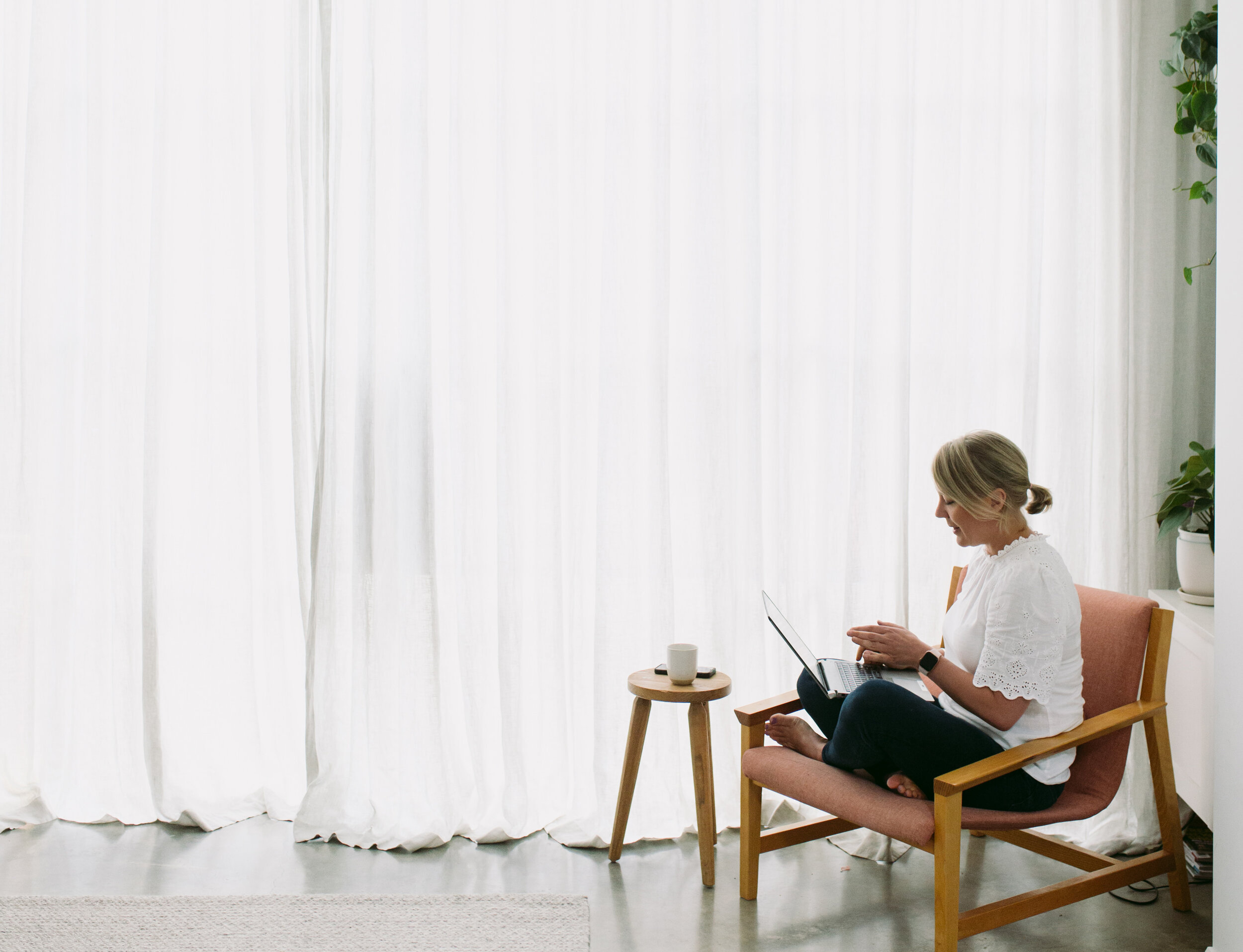 A woman sitting cross-legged on a pink armchair, working on a laptop, with a white curtain backdrop, a small wooden side table with a mug, and some potted plants nearby.