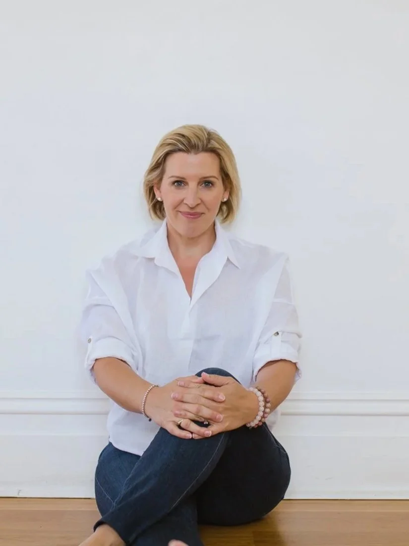 A woman with blonde hair sitting on the floor against a white wall, wearing a white shirt and dark jeans, with her hands clasped on her knee.