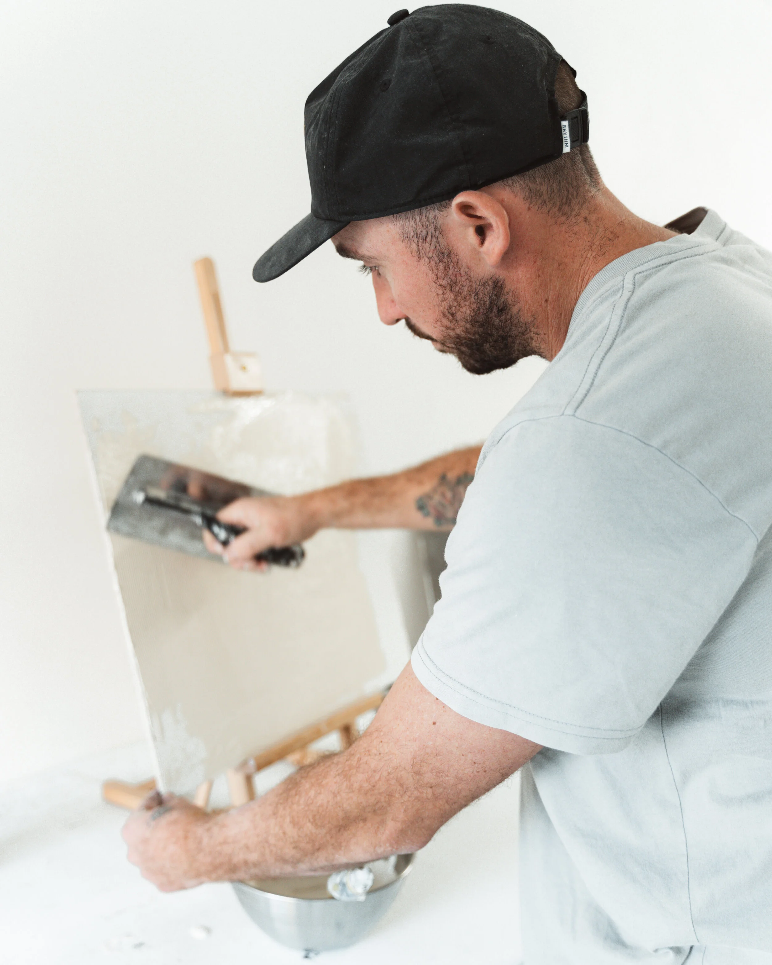 Man using spatula on canvas with plaster, wearing cap