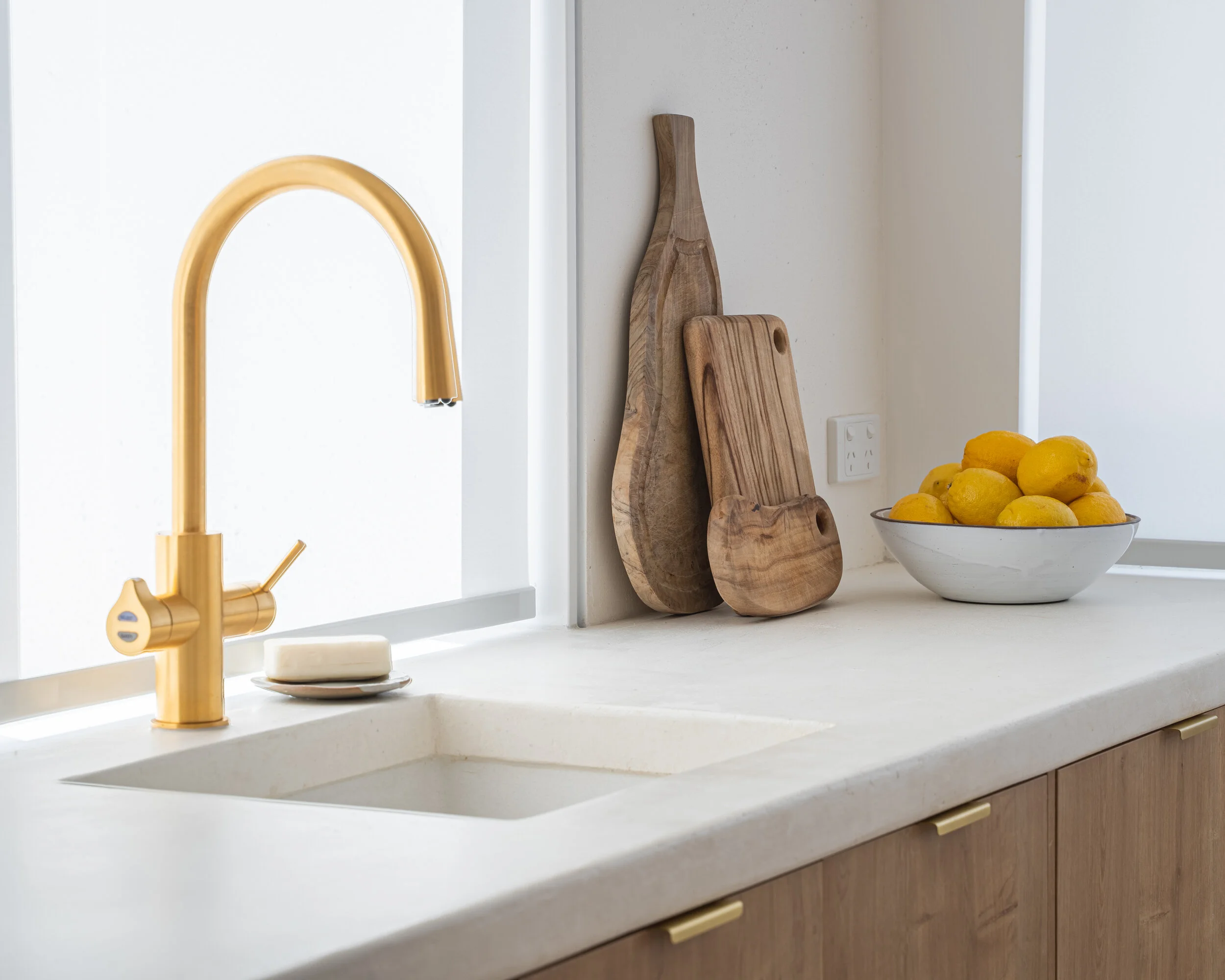 Modern kitchen countertop with gold faucet, white sink, wooden cutting boards, and a bowl of lemons.