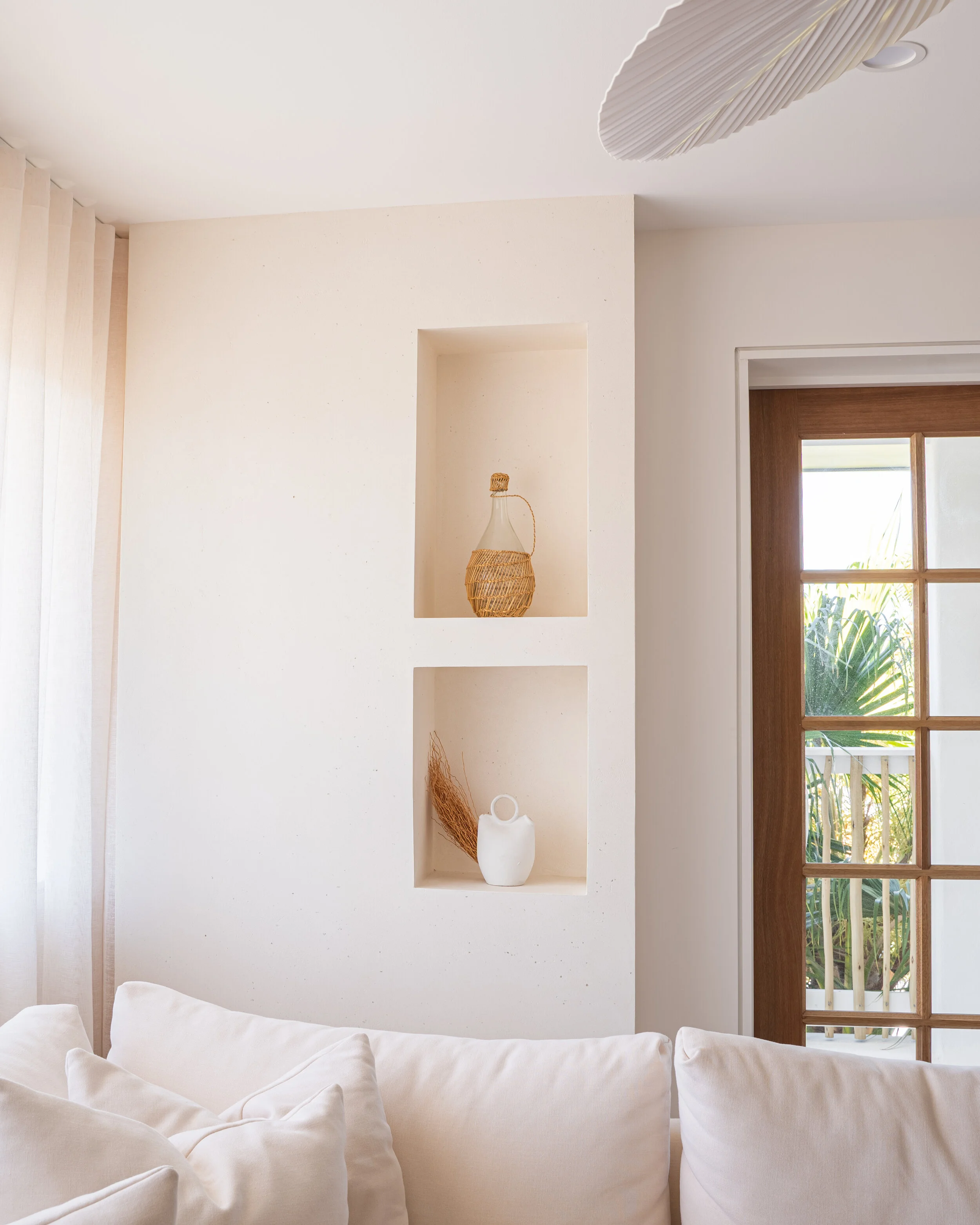 Minimalist living room with built-in wall shelves, wicker-wrapped bottle, white ceramic jug, soft white sofa, sheer curtains, and glass door leading outside.