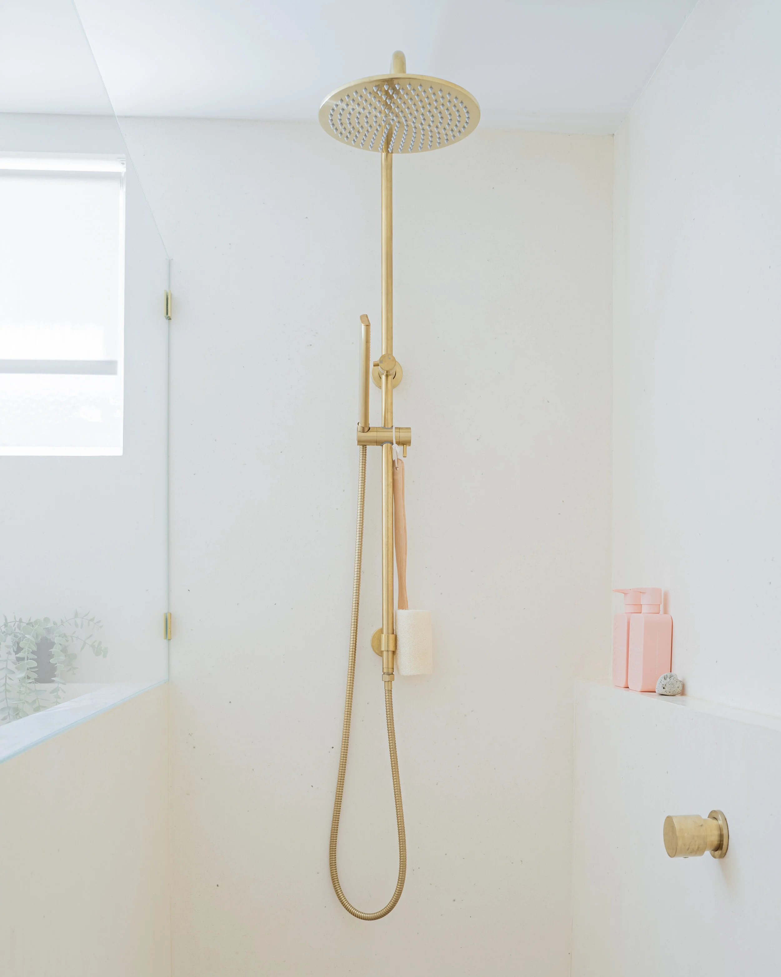 Minimalist bathroom with a gold-colored rain showerhead, hand shower, glass enclosure, and small shelf with pink soap dispenser and stone.