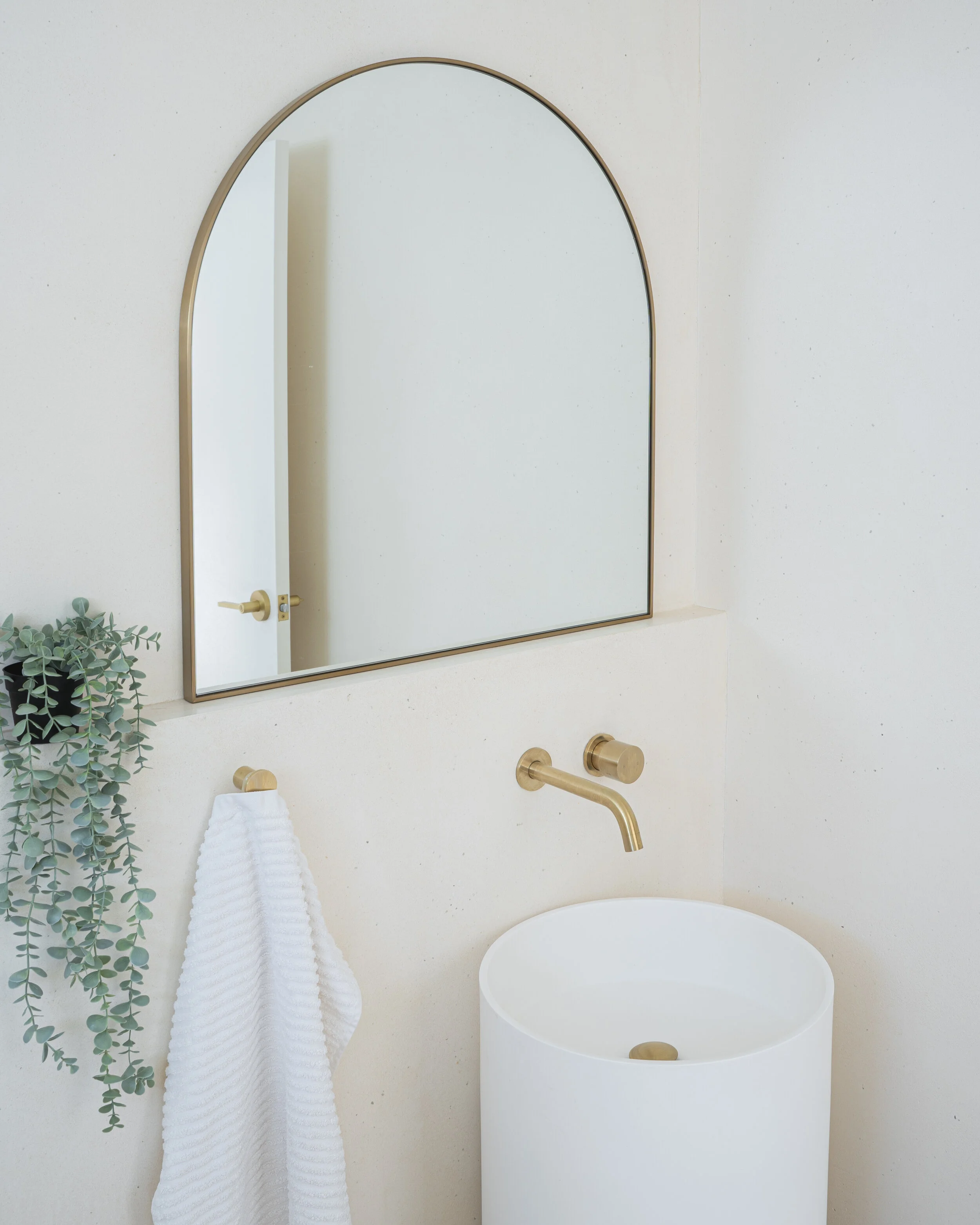 Modern bathroom with an arched mirror, gold faucet, white vessel sink, eucalyptus plant, and white towel.