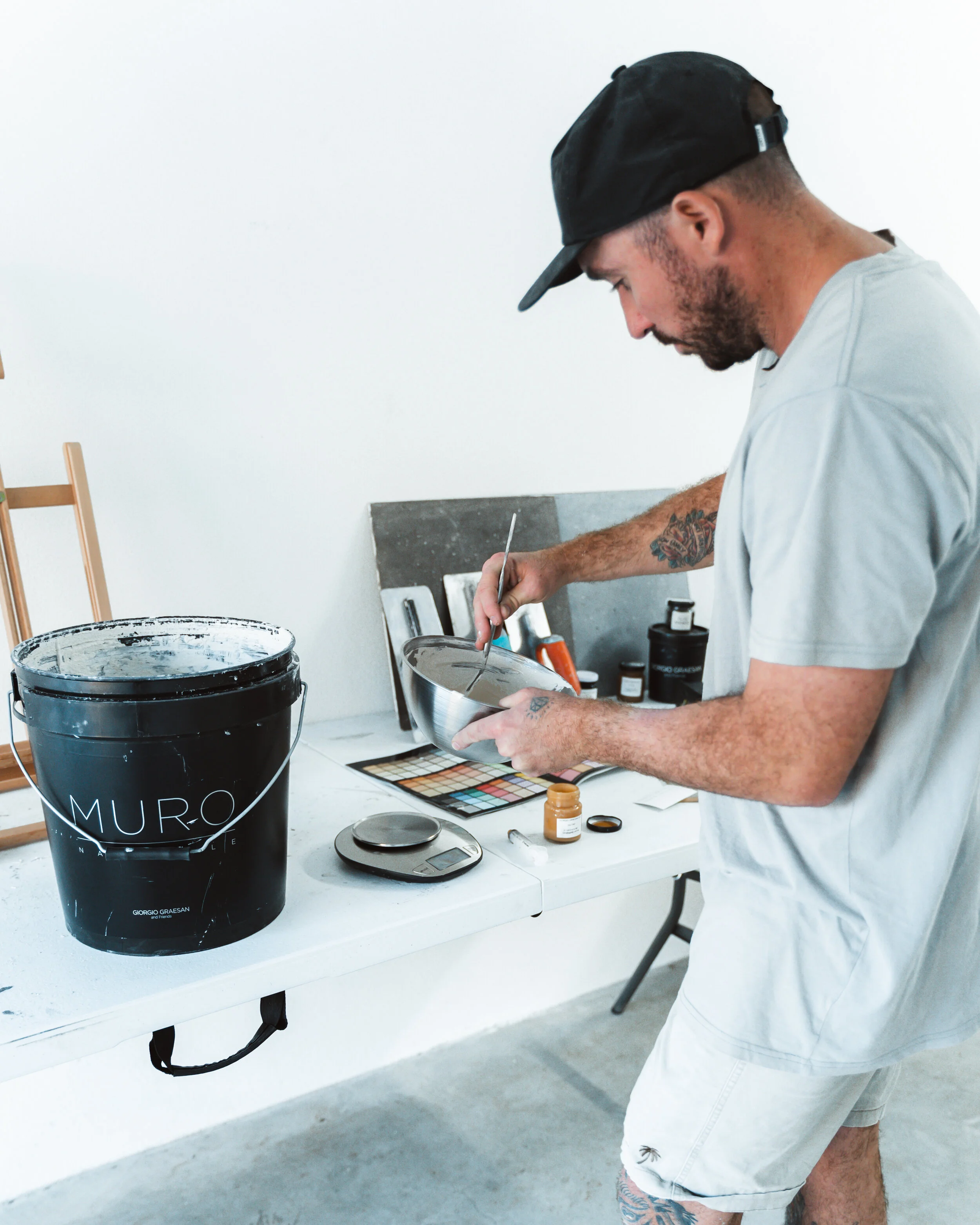 Man mixing paint at a table with various art supplies, including a large black bucket labeled "MURO" and color samples.