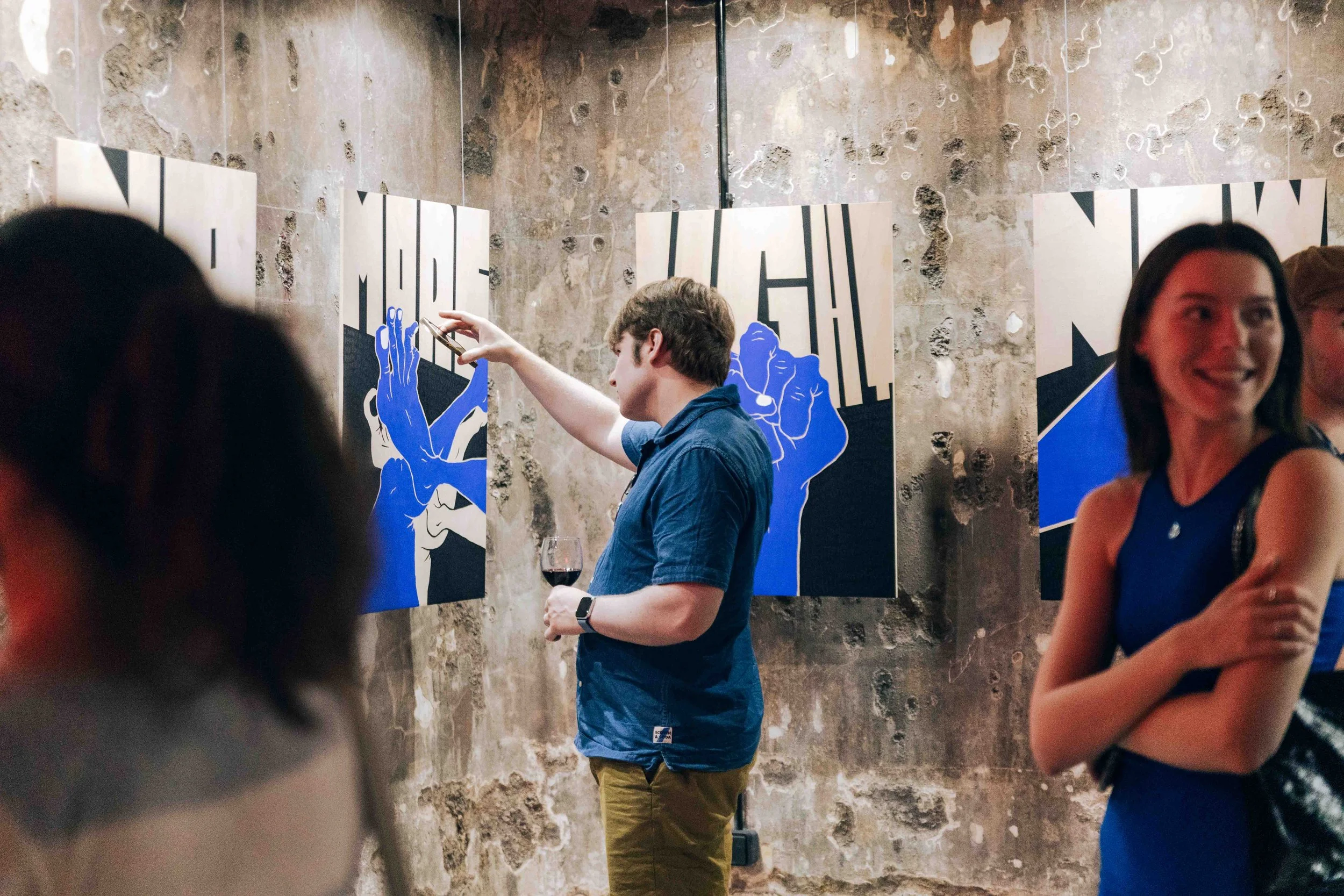 People attending an art gallery or exhibition, with one person in a blue shirt taking a photo of artwork on a textured wall, and a woman smiling in the foreground.
