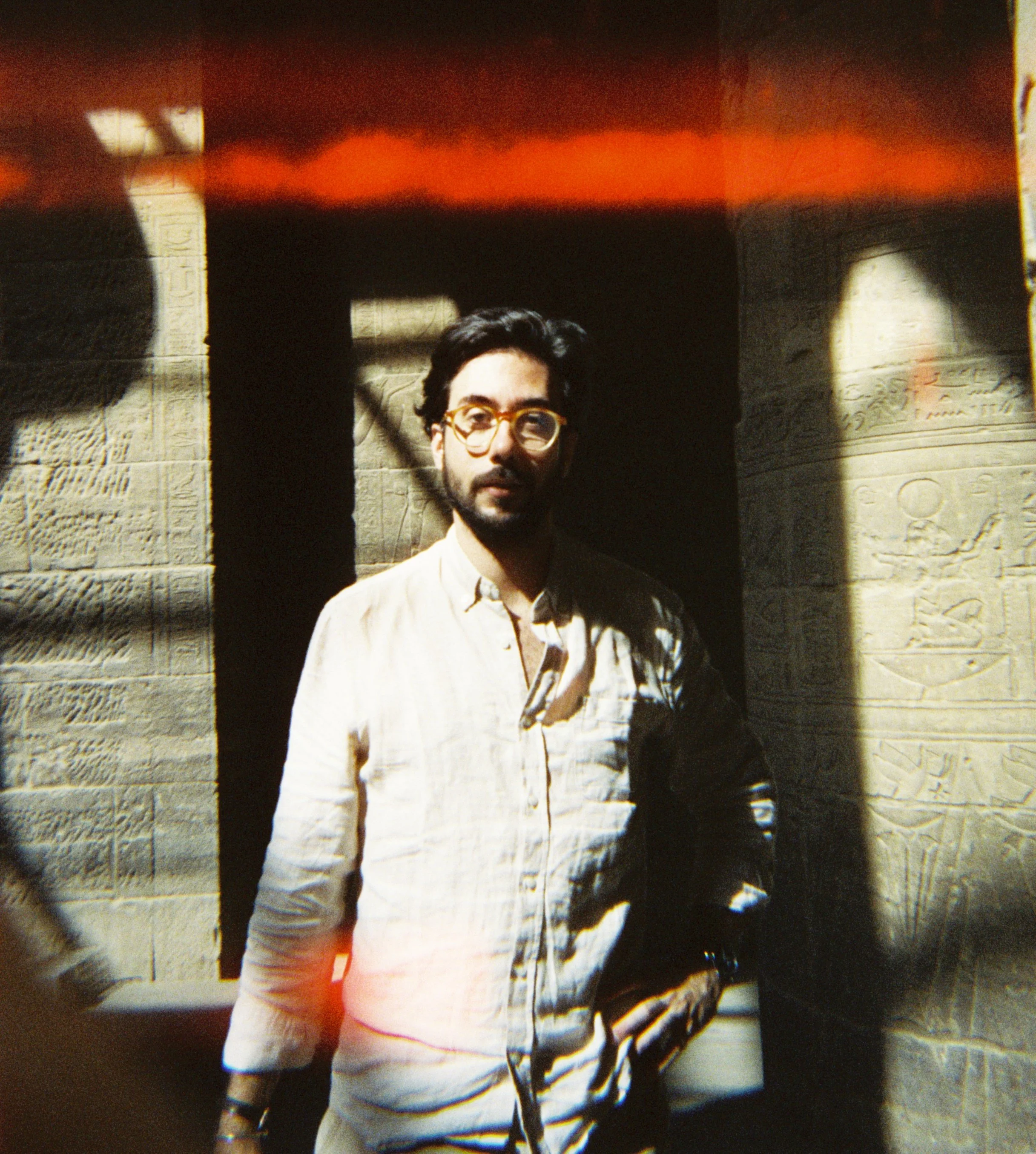 A man with glasses and dark wavy hair stands in front of an ancient Egyptian wall with hieroglyphics, partly in sunlight and shadow.