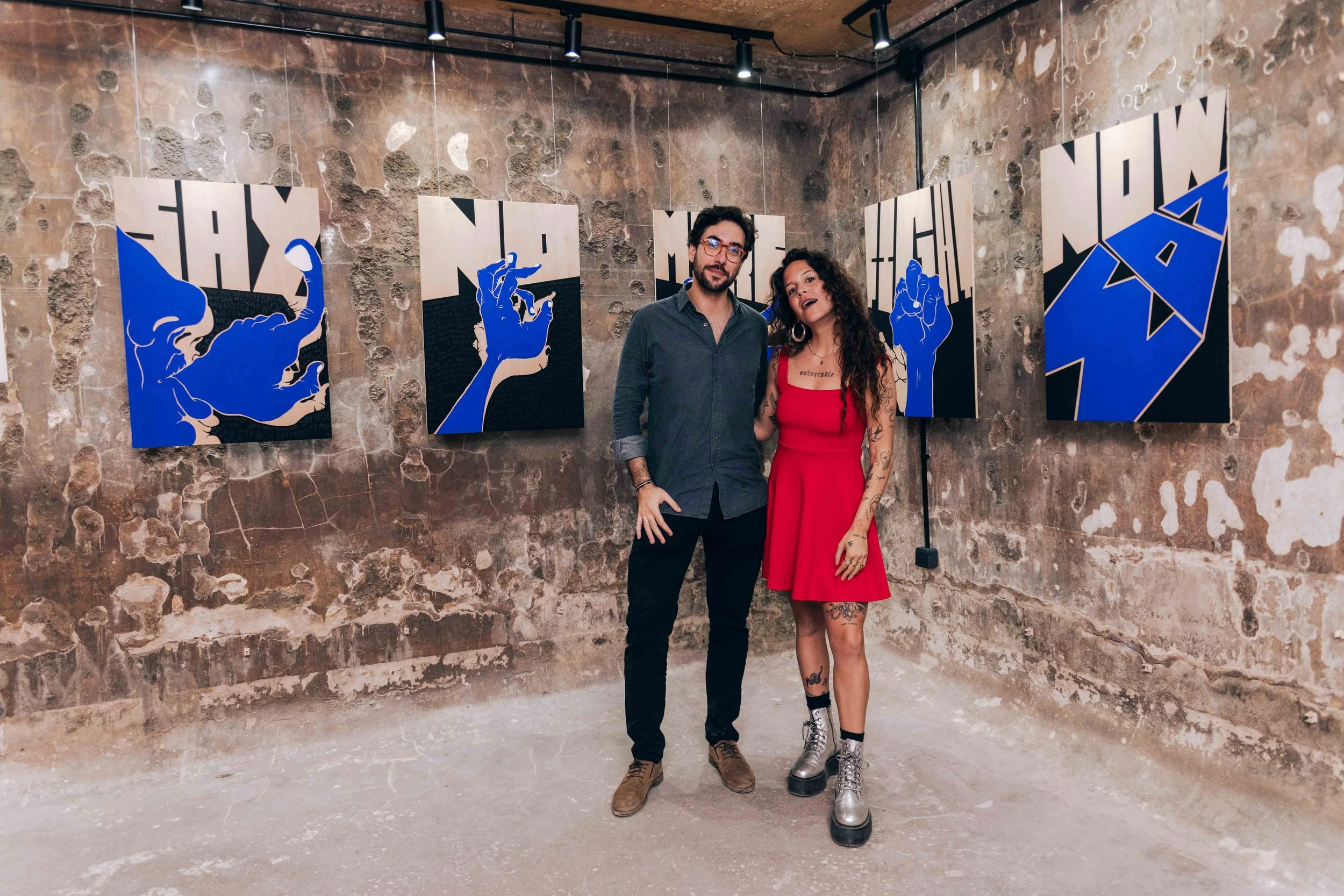 A man and woman stand close together in an art gallery with stone walls and abstract blue and black artwork. The man wears glasses, a dark shirt, and pants, while the woman has dark curly hair, tattoos on her arms and legs, and wears a red dress with combat boots.