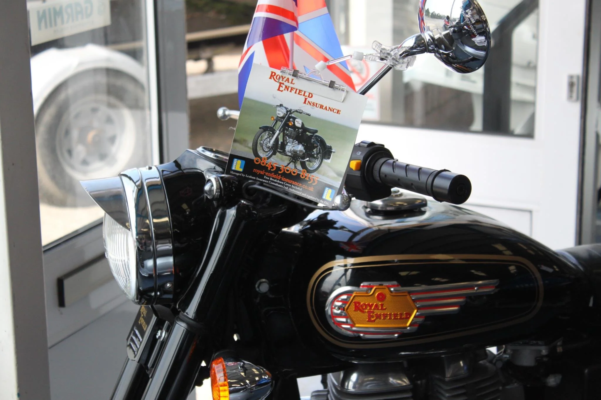 Close-up of a black Royal Enfield motorcycle displayed inside a store, with a flyer advertising Royal Enfield insurance placed on the handlebar.