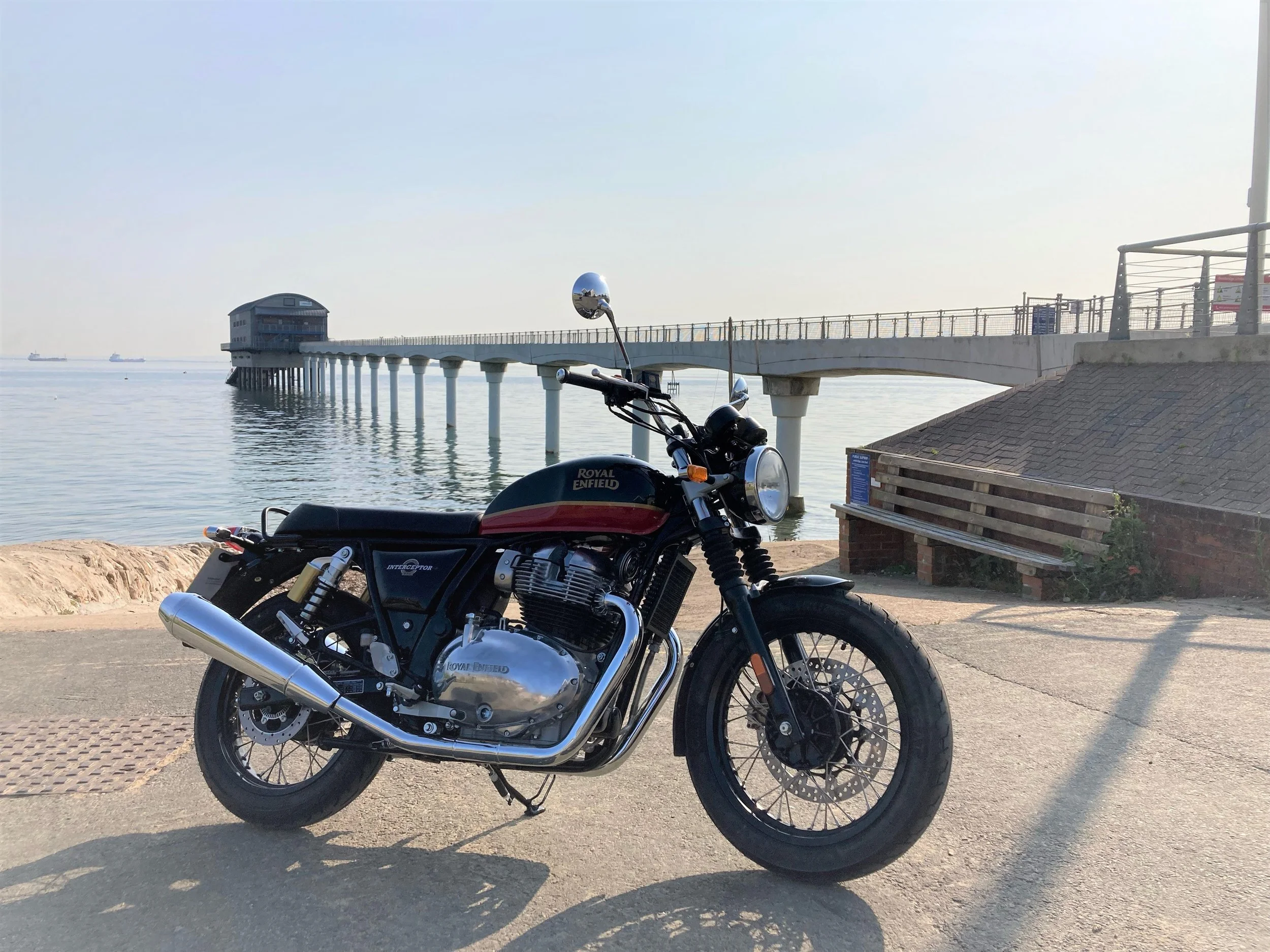 A black and red Royal Enfield Interceptor motorcycle parked near the water with a pier extending into the ocean in the background.
