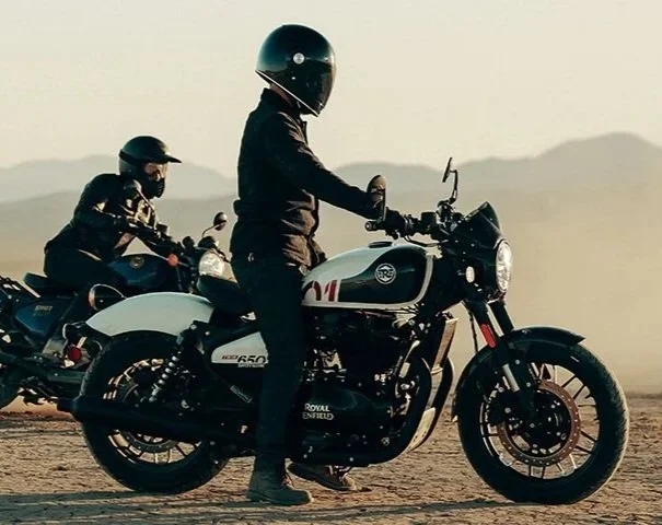 Two people riding Royal Enfield motorcycles in a desert landscape during sunset or sunrise.