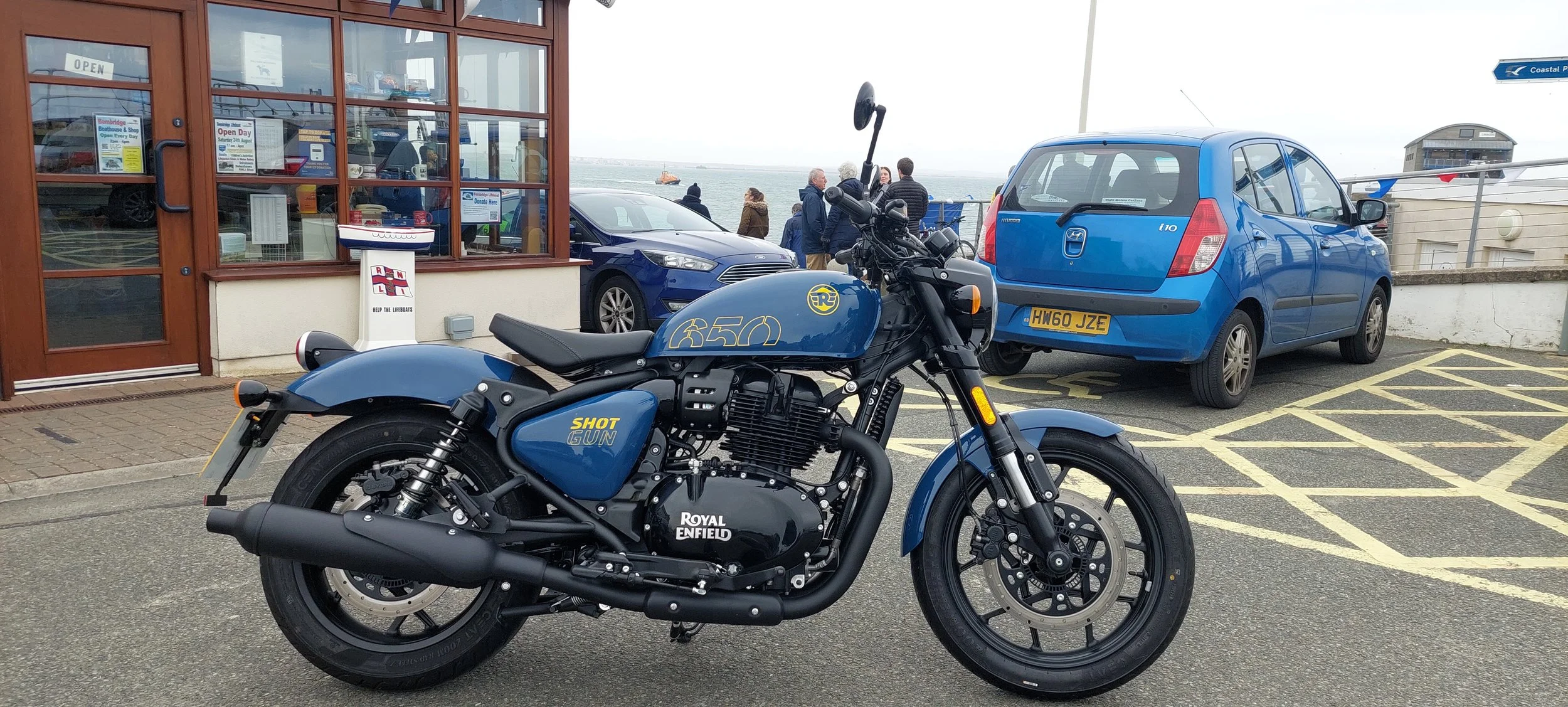 Blue Royal Enfield Shotgun 650 motorcycle parked in a seaside parking lot with cars and a group of people near the shoreline.