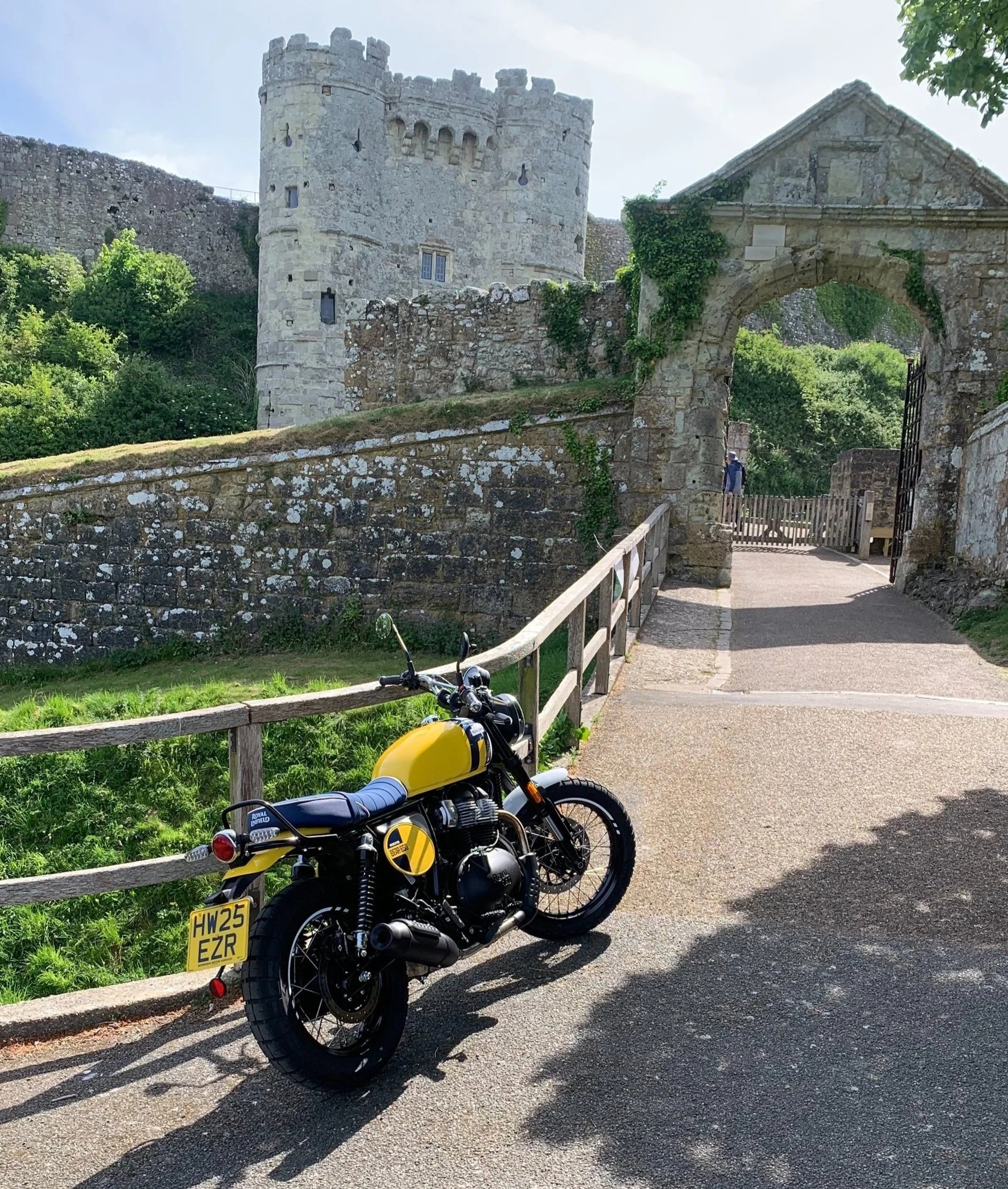 A yellow and black Royal Enfield Bear 650 motorcycle parked on a path in front of an ancient stone Carisbrooke castle on the Isle of Wight with a tower, archway, and greenery.
