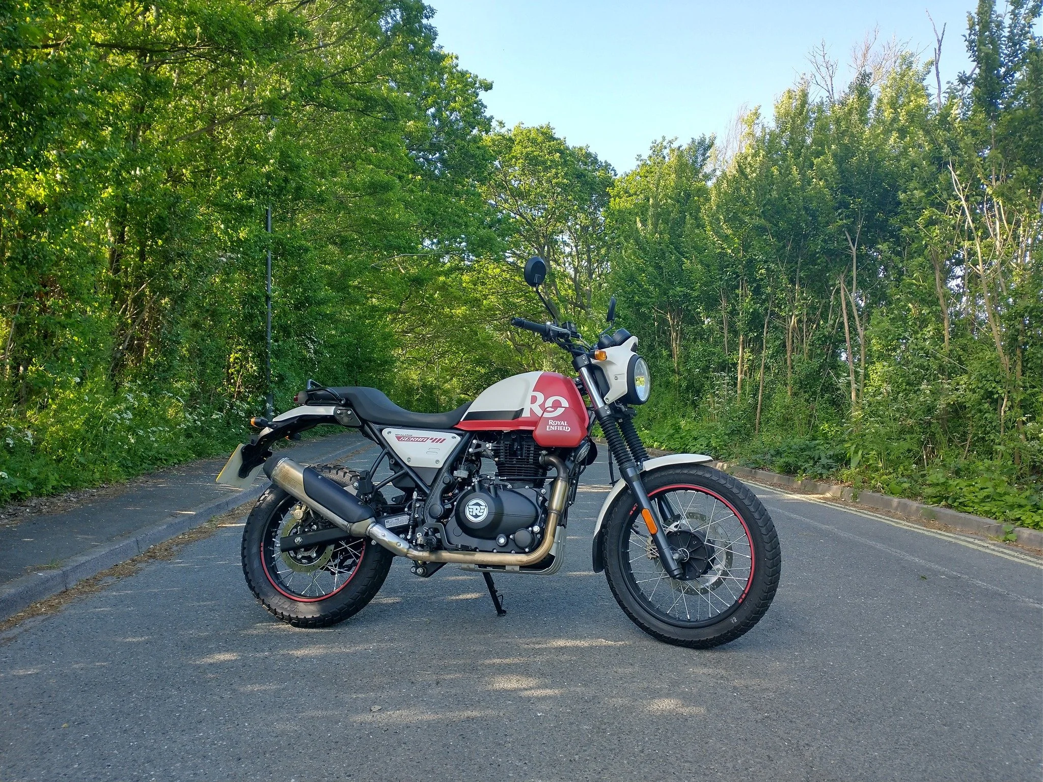 A Royal Enfield motorcycle parked on an asphalt road surrounded by green trees on a sunny day.