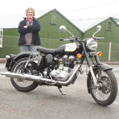 A smiling man with gray, curly hair wearing a black and gray jacket and jeans standing with arms crossed next to a black and white Royal Enfield motorcycle on a street.