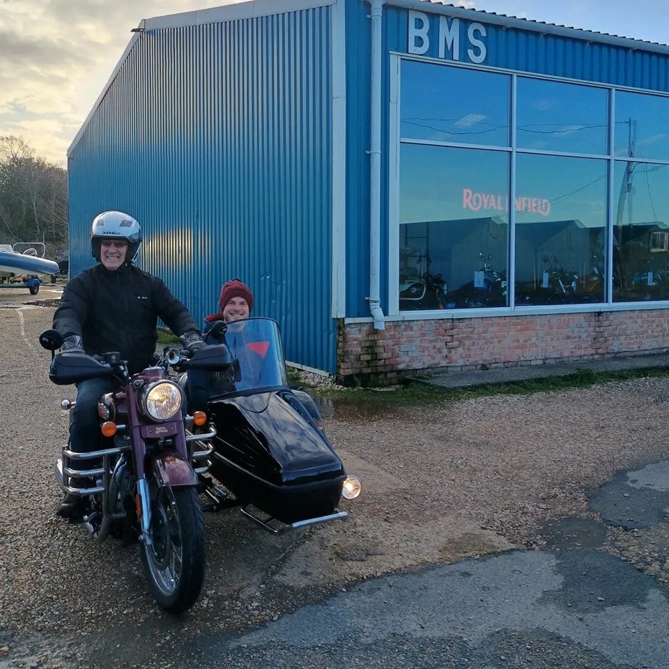 A person riding a Royal Enfield motorcycle with a sidecar, smiling, wearing a black jacket and helmet, parked in front of a blue building with large windows, and a woman sitting inside the sidecar, wearing a red beanie, smiling.