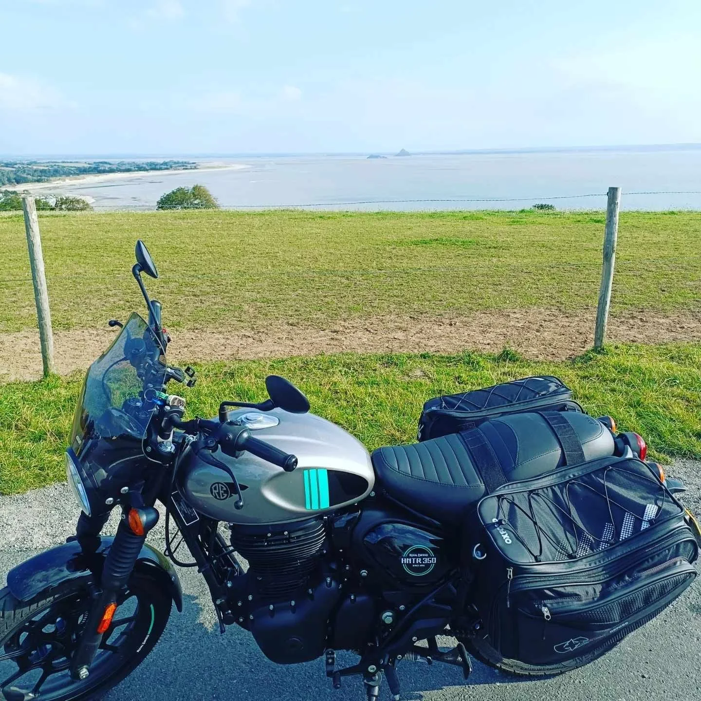 A Royal Enfield motorcycle parked on a paved area near a grassy field with a view of water and distant shoreline beyond.