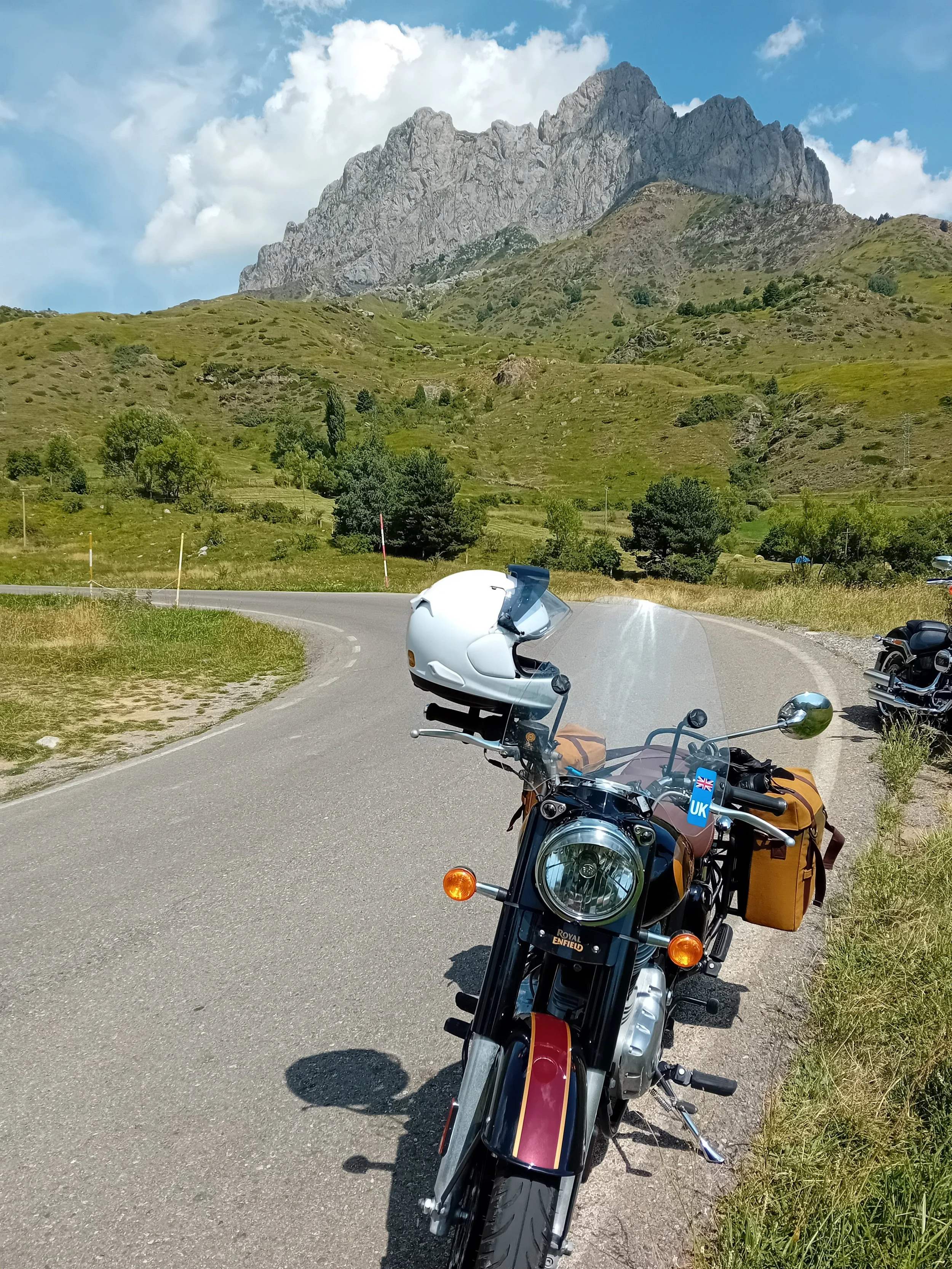 A Royal Enfield motorcycle with a white helmet on it parked on a winding road surrounded by grassy hills and green trees, with a mountain in the background under a partly cloudy sky.