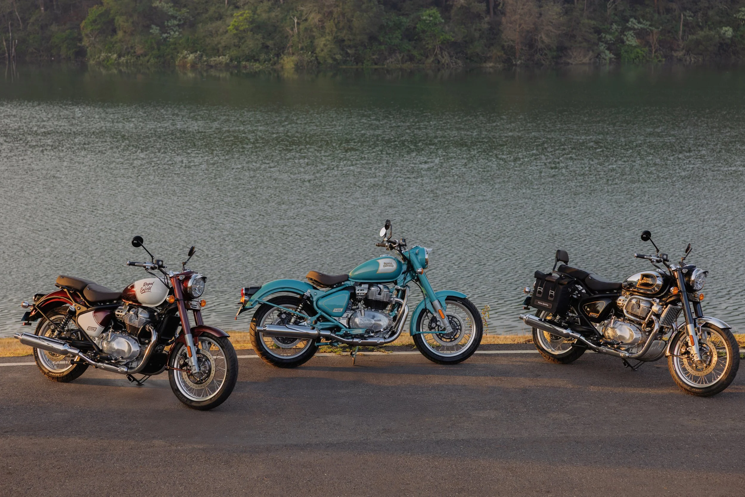 Three Royal Enfield Classic 650 motorcycles parked on the side of a road near a body of water with a tree-lined shoreline in the background.