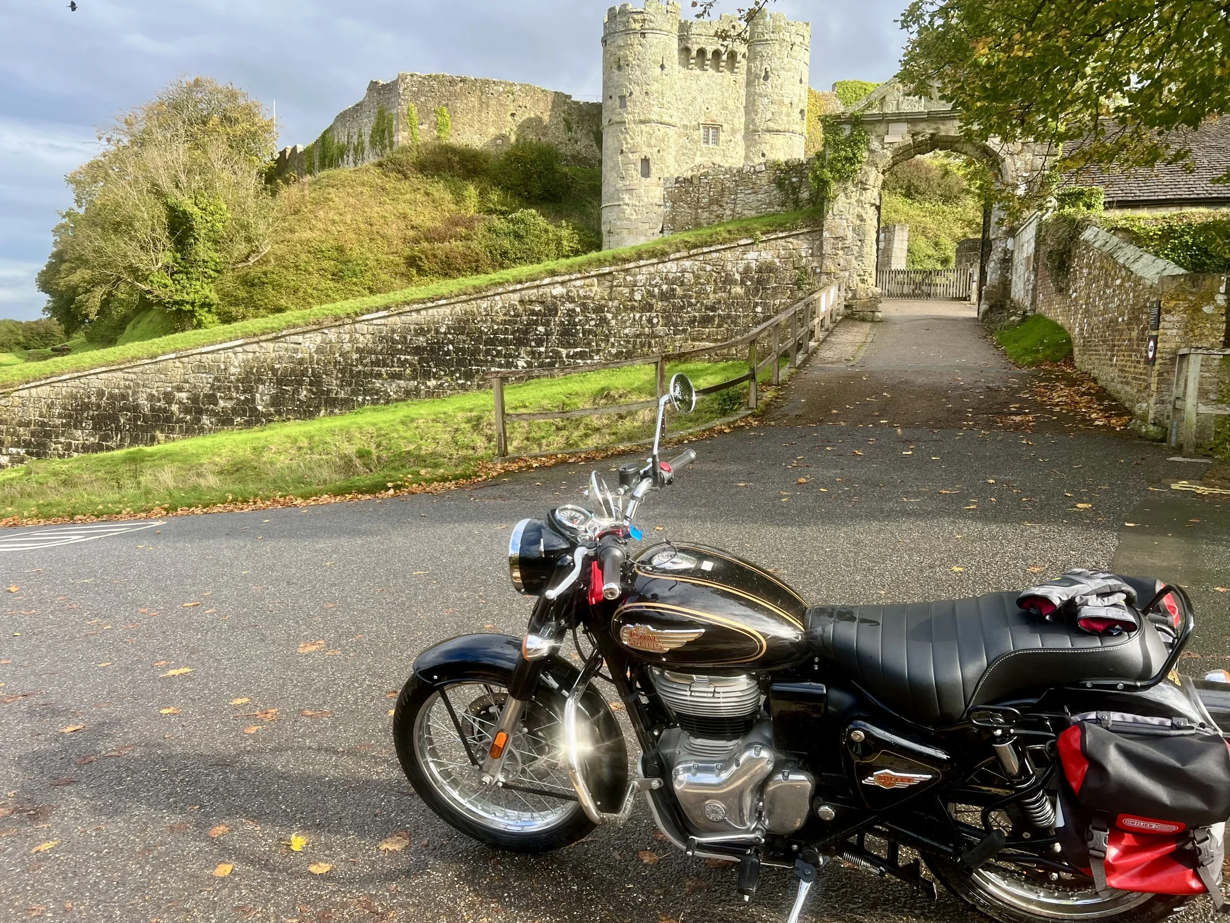 A black Royal Enfield motorcycle parked in front of a historic castle with stone walls, a tower, and an arched entrance, surrounded by trees with fall foliage.