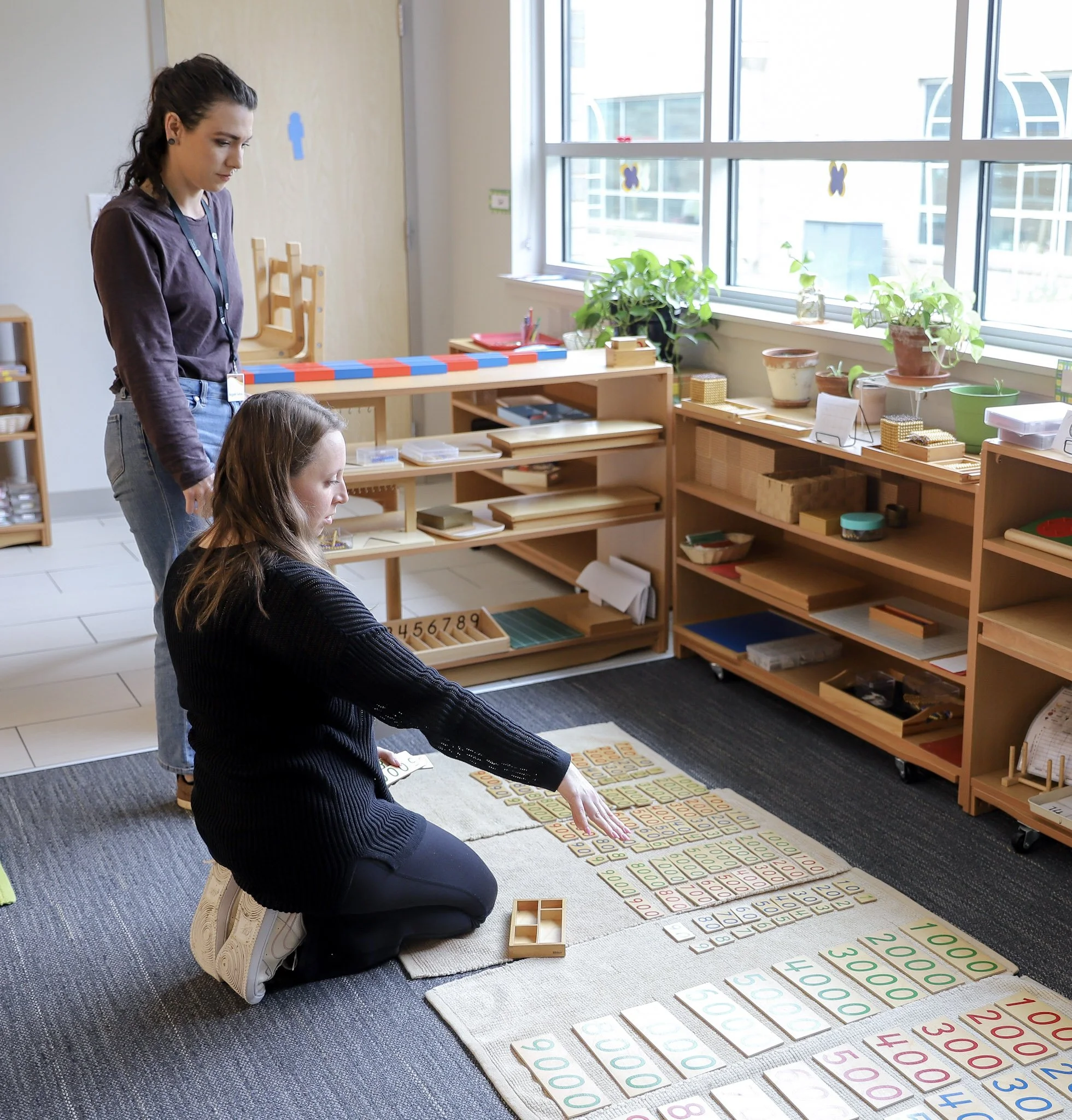 Two women are working on a Montessori math activity with number cards and tiles, in a classroom with shelves, plants, and large windows.