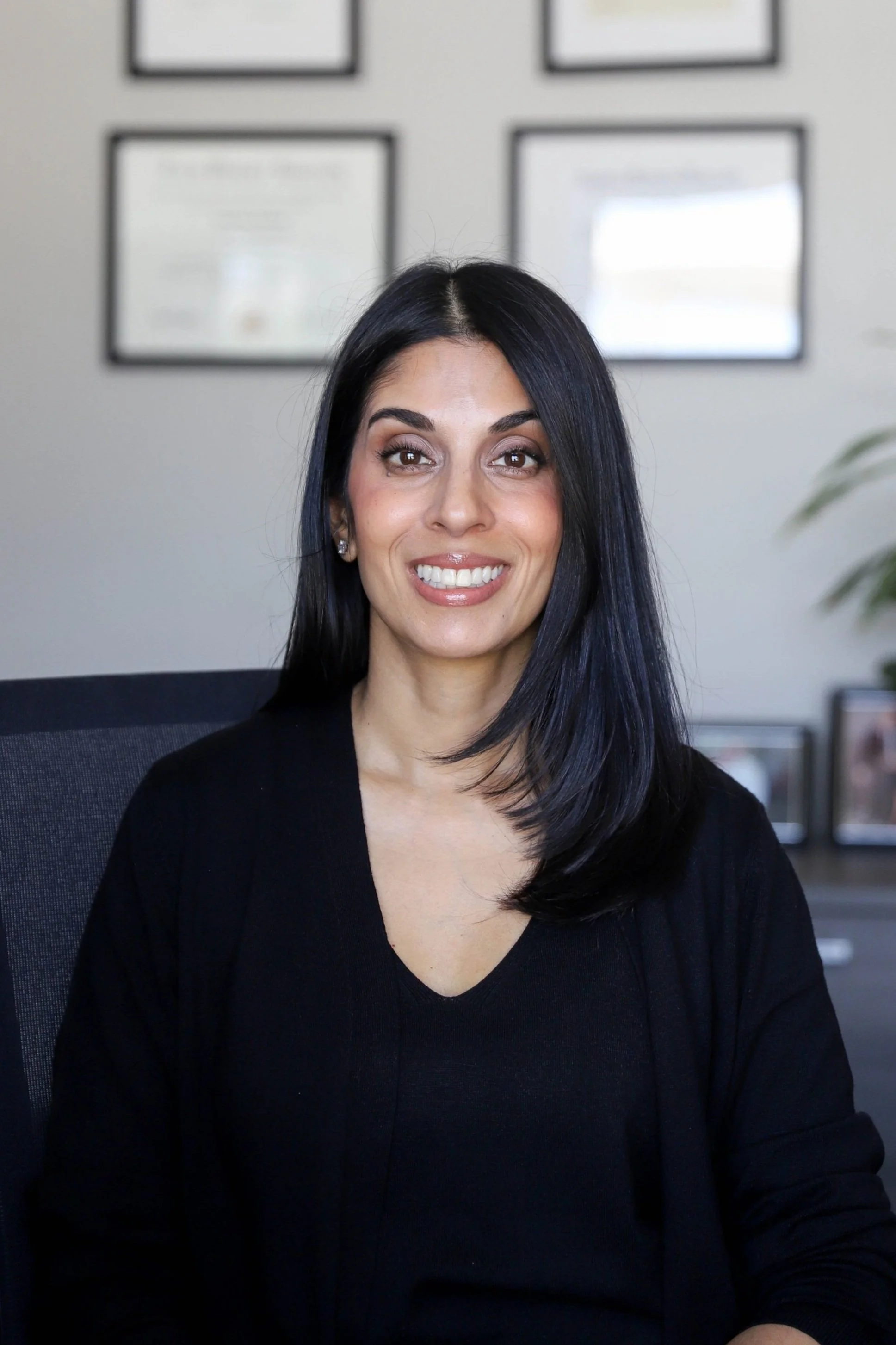 A woman with long black hair and a black top smiling at the camera, sitting in an office with framed certificates on the wall behind her.