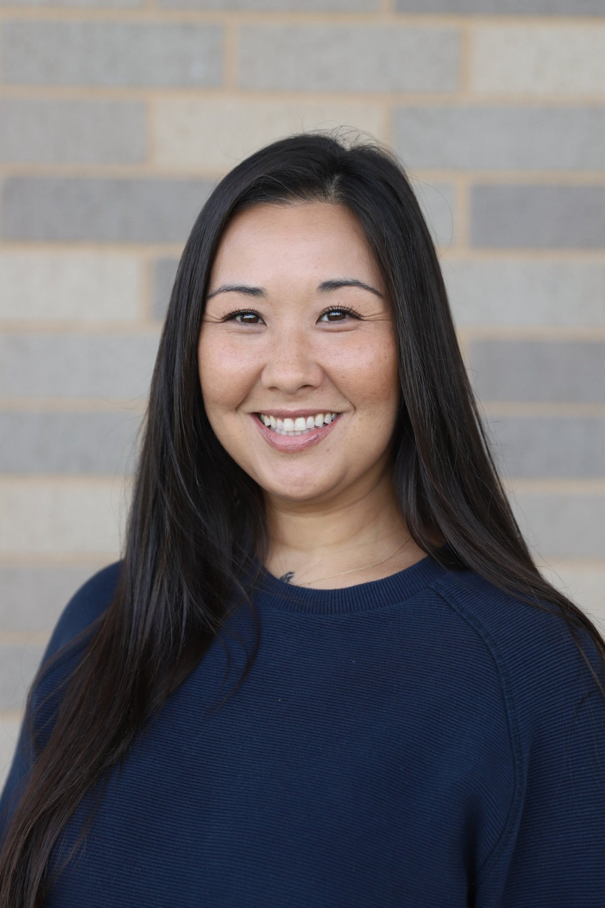A smiling woman with long dark hair wearing a navy blue top standing against a brick wall background.