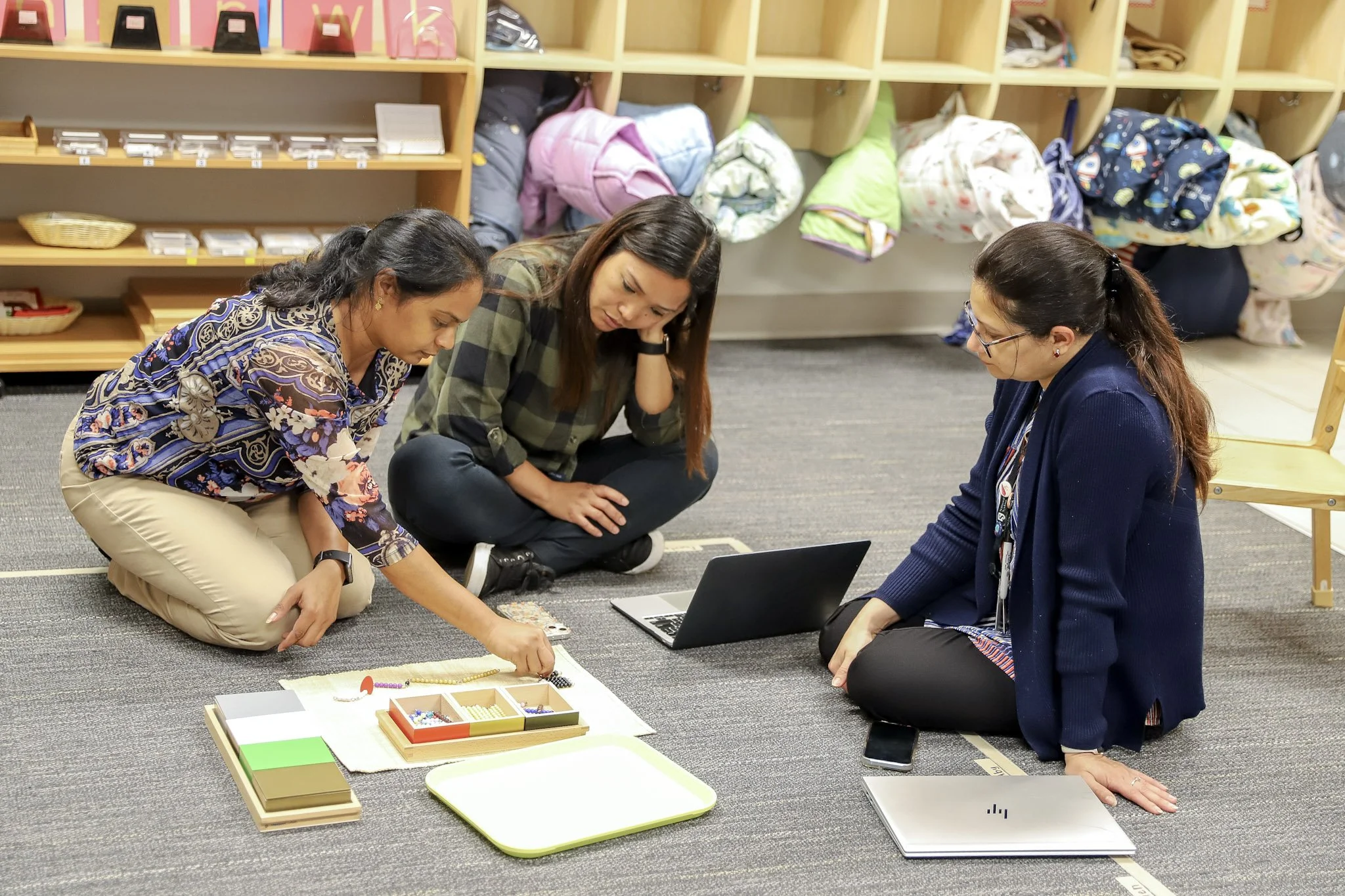 Three adult learners sitting on the floor in a Montessori classroom with cubby shelves, examining Montessori math beads.