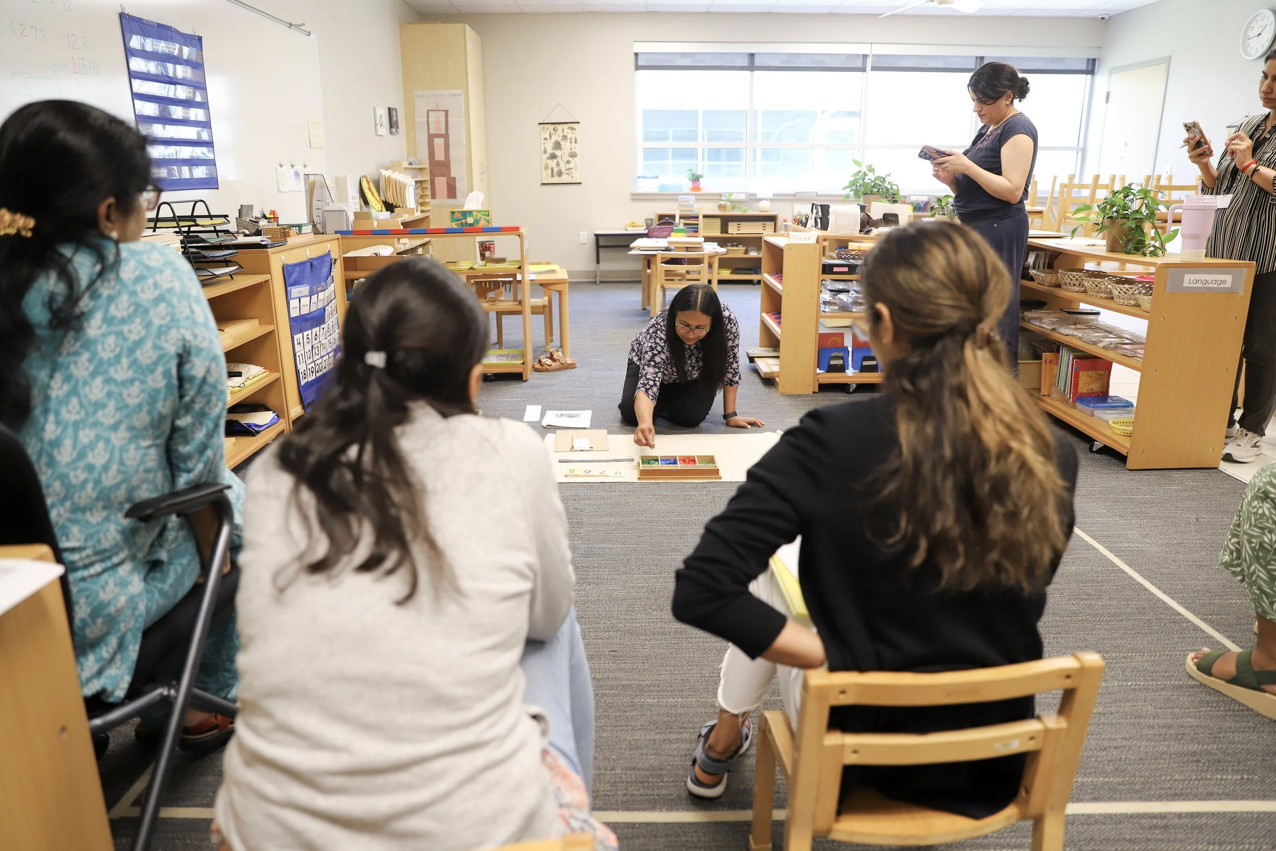 Teacher and students in a classroom participating in a Montessori work on the floor, with Montessori shelves, windows, and students taking notes in the background.