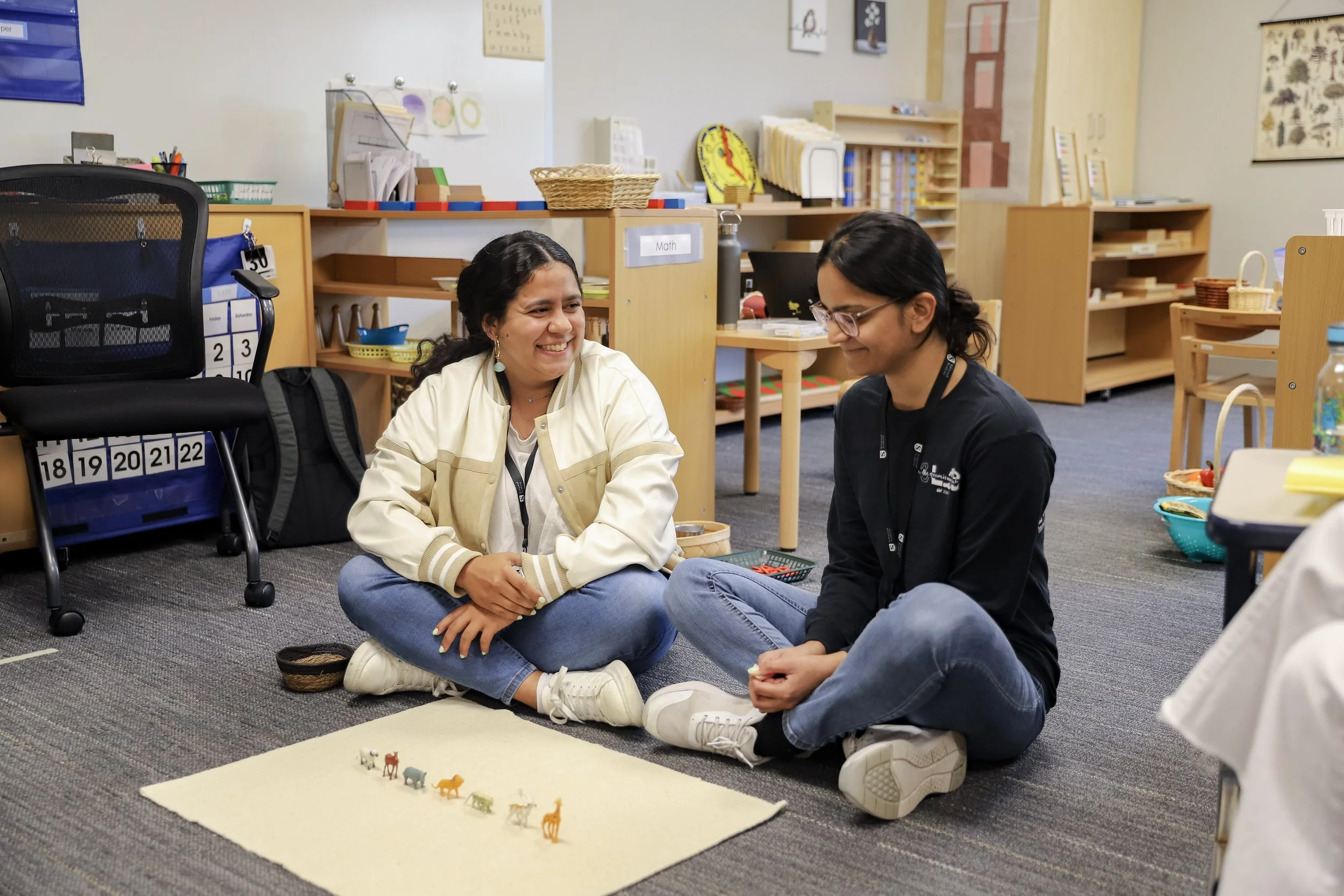 Two adult leaners sitting on the floor of a classroom, engaging in a Montessori language lesson with small animal figurines on a Montessori rug. They are smiling and appear to be in a friendly conversation.