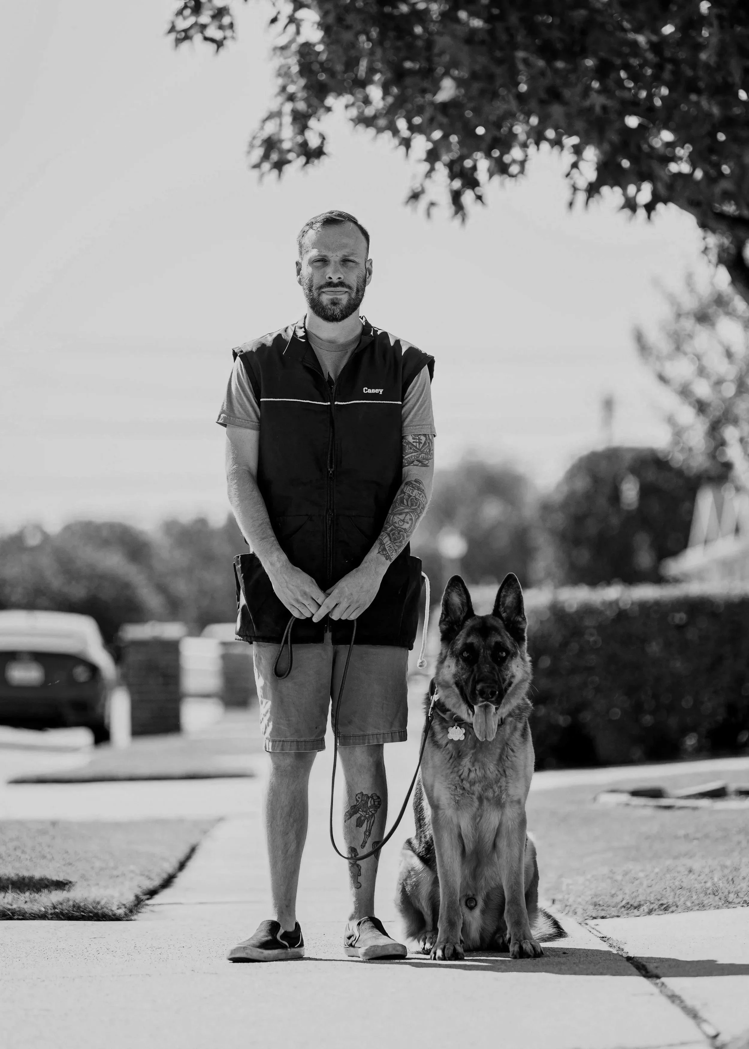A man standing outdoors in Austin Texas on a sidewalk holding a leash attached to a German Shepherd dog sitting beside him, with trees and cars in the background. Legion Dog Training